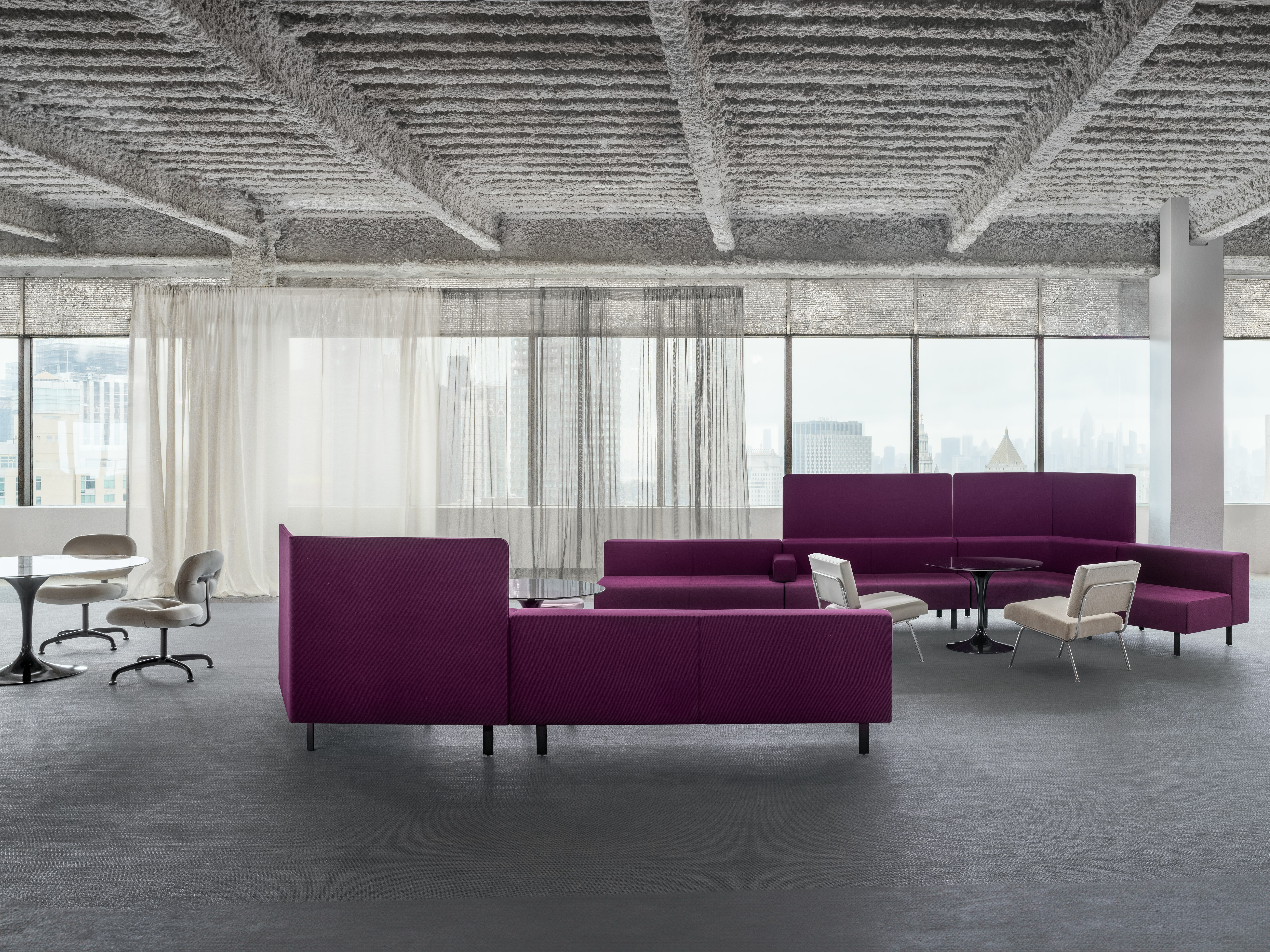 A group of young seating is sitting in an open office space with a long group of windows in the background. Sheer drapes are covering some of the windows.