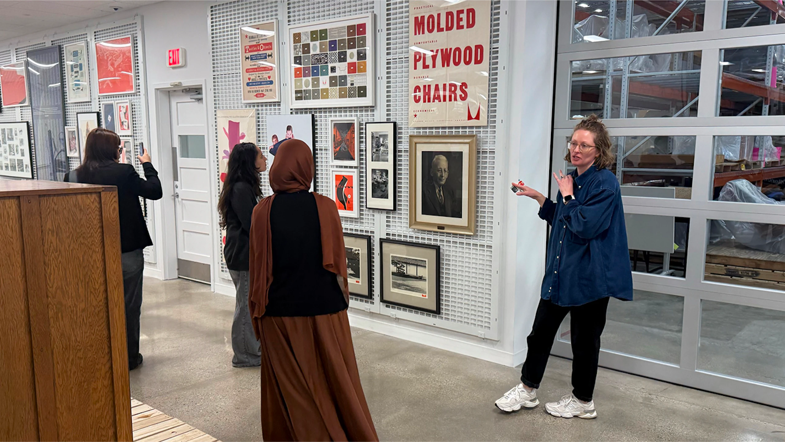 A woman in a blue shirt and black pants gives a tour of the MillerKnoll Archives to a small group of people. Many historic art pieces can be seen hanging on the wall next to a warehouse door.