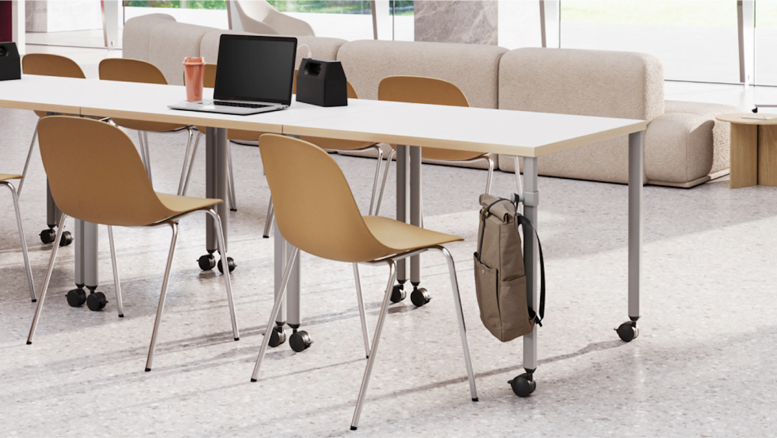 Modern space with long white table, tan rolling chairs, and a laptop. A backpack hangs on the table's hook. Soft seating is visible in the background.