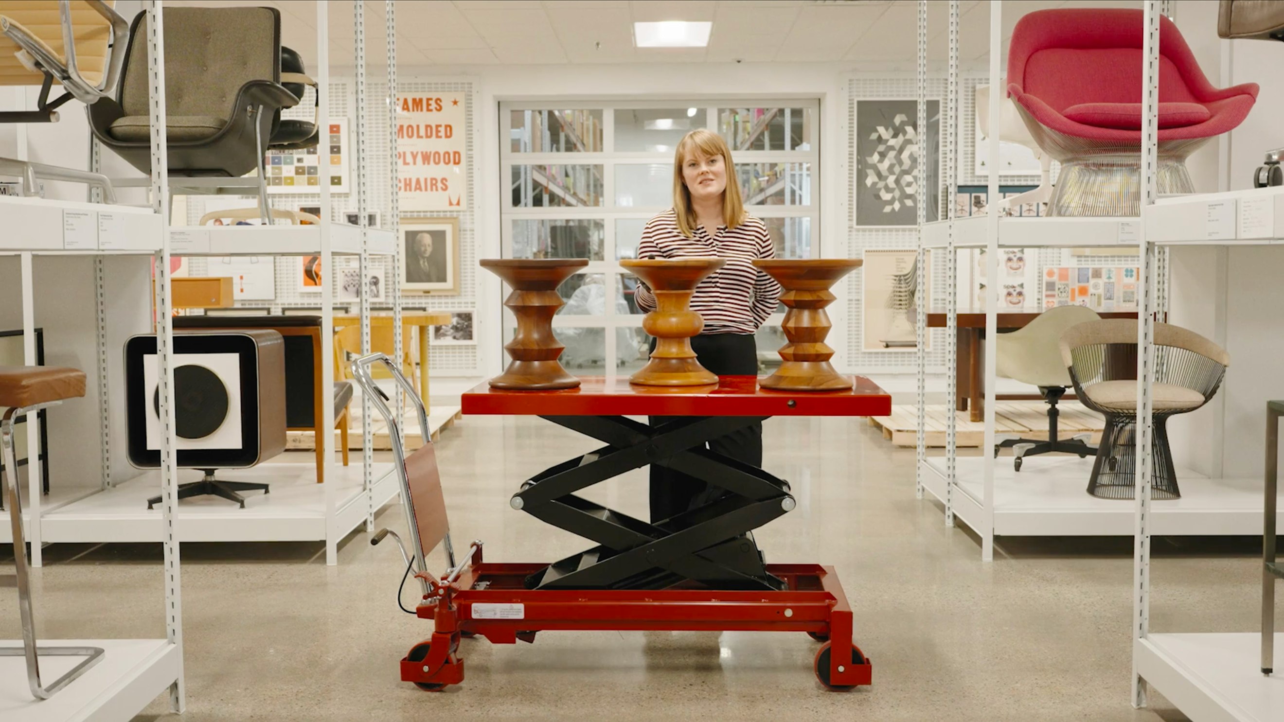 Stine Aas standing behind three Eames Turned Stools on a red scissor lift among the racks at the MillerKnoll Archives.