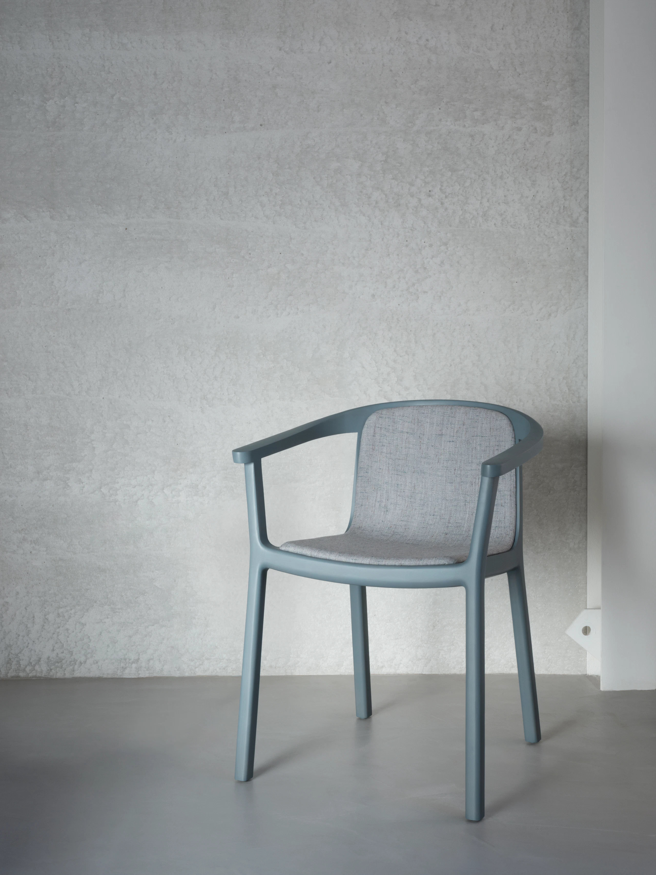 Lijn Armchair in with Nordic Blue frame and an upholstered seat in back in a light blue textile, viewed at an angle against a grey wall and concrete floor.