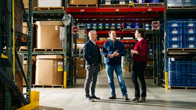 Three people engaged in conversation inside a warehouse.