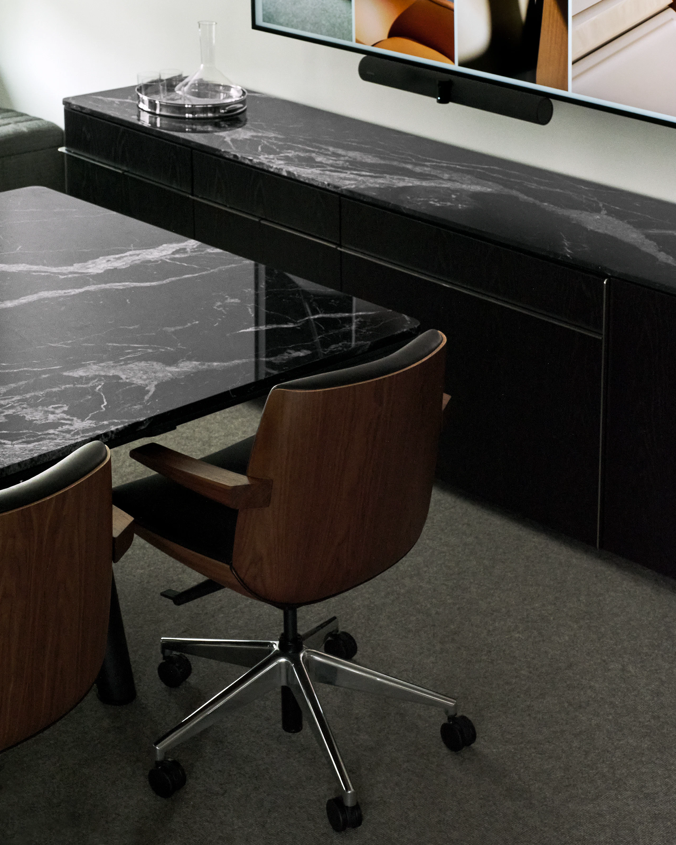 Two low back Clamshell Conference Chairs in Walnut upholstered with black lather beside an Ascent Conference Table and Credenza, both with Nero Marquina Marble tops, viewed from behind at an angle.