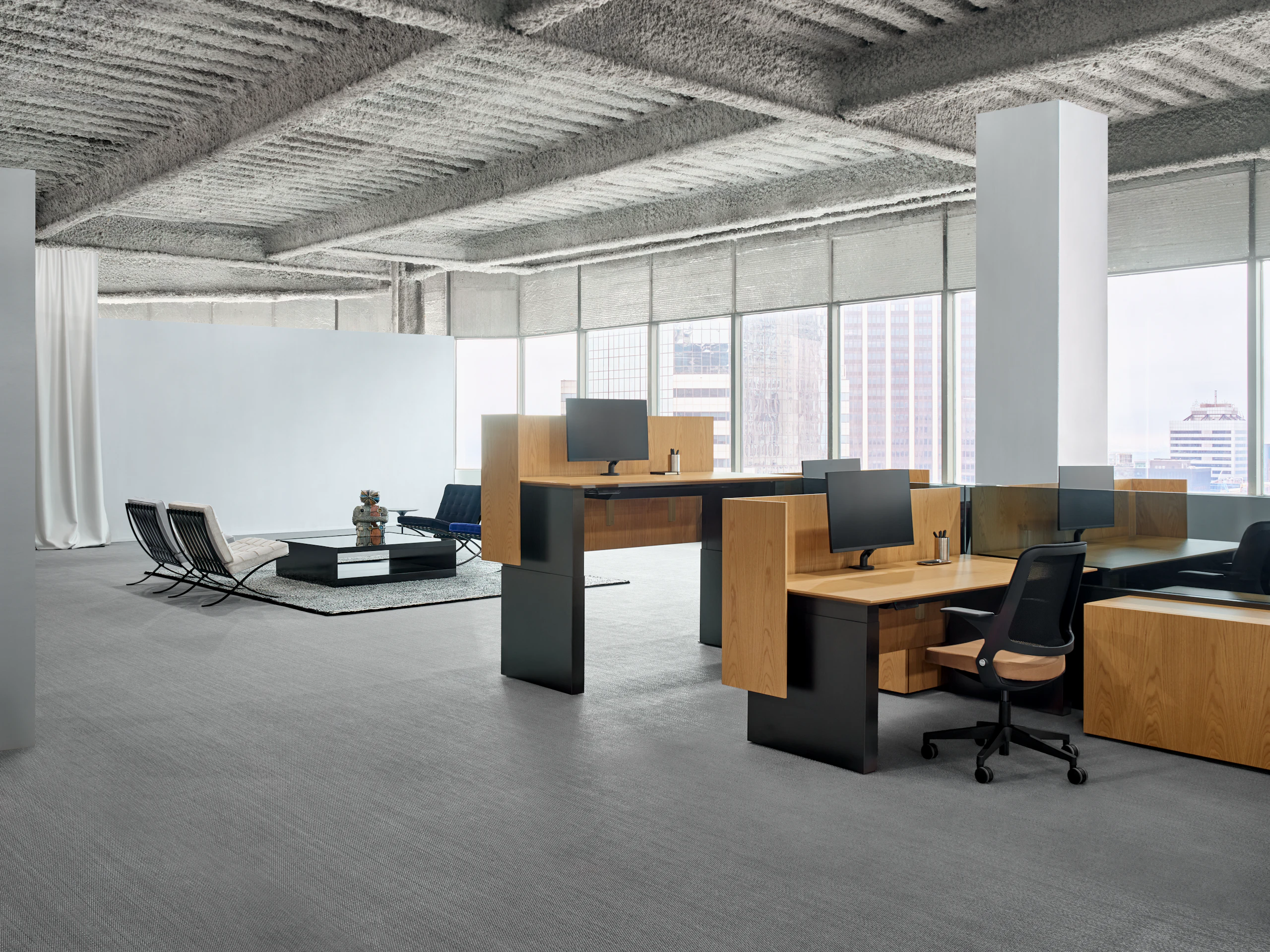 A group of workstations with height adjustable work tables are in front of a lounge area with four chairs.