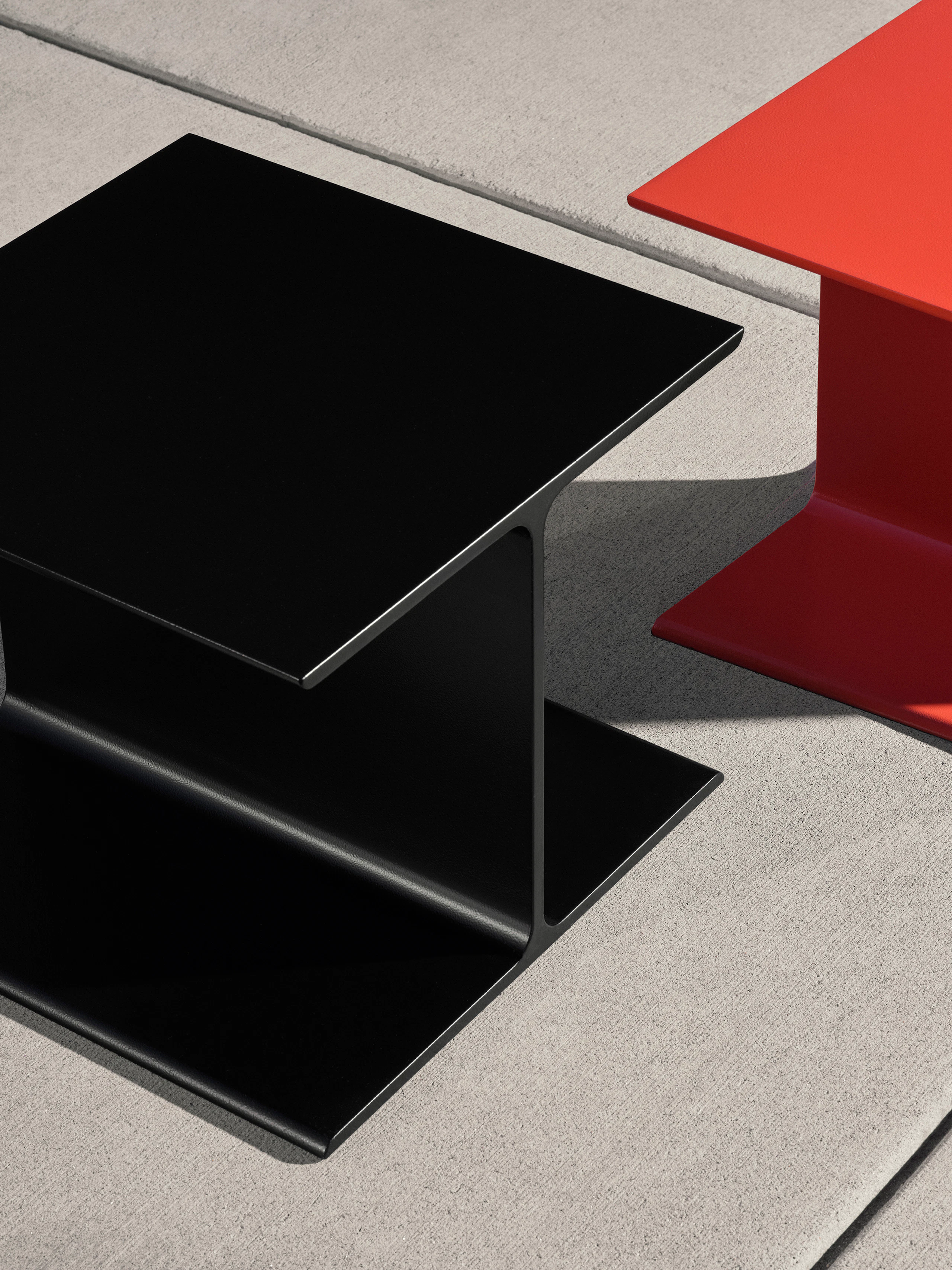 Partial view of two I Beam Side Tables designed by Ward Bennett, one in black positioned slightly in front of one in International Orange, viewed from above at an angle on a concrete floor.