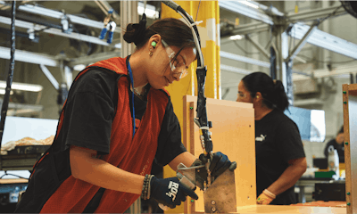 A woman wearing safety glasses, a red vest, a black shirt, and black gloves works on a furniture assembly line. Pieces of wood with green dowels can be seen at her station.