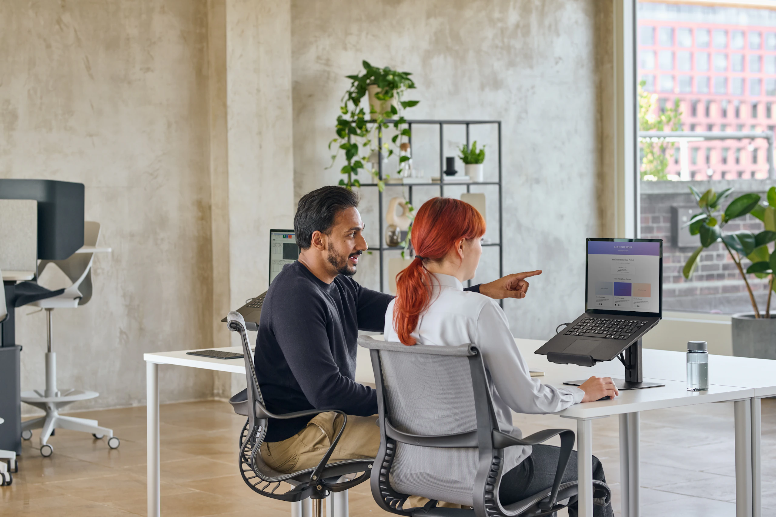 Person a with red hair using black Lana laptop stand while person b with black hair pointing at the laptop in a work environment.