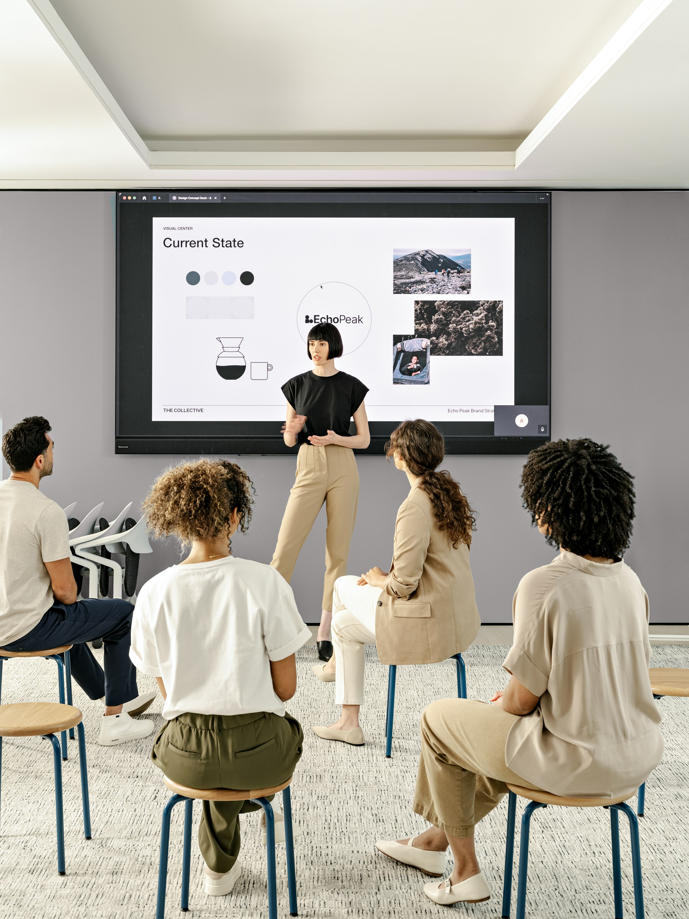 A woman standing in front of a screen giving a presentation to an audience of three women and one man seated on stools.