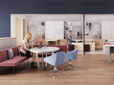 A modern workspace with wooden flooring, featuring an individual seated at a round table, along with a person working at a desk behind glass, and contemporary furniture, including red and blue chairs.