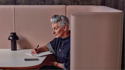 A woman with short grey hair sitting in a light pink booth at a white table while writing in a notebook.