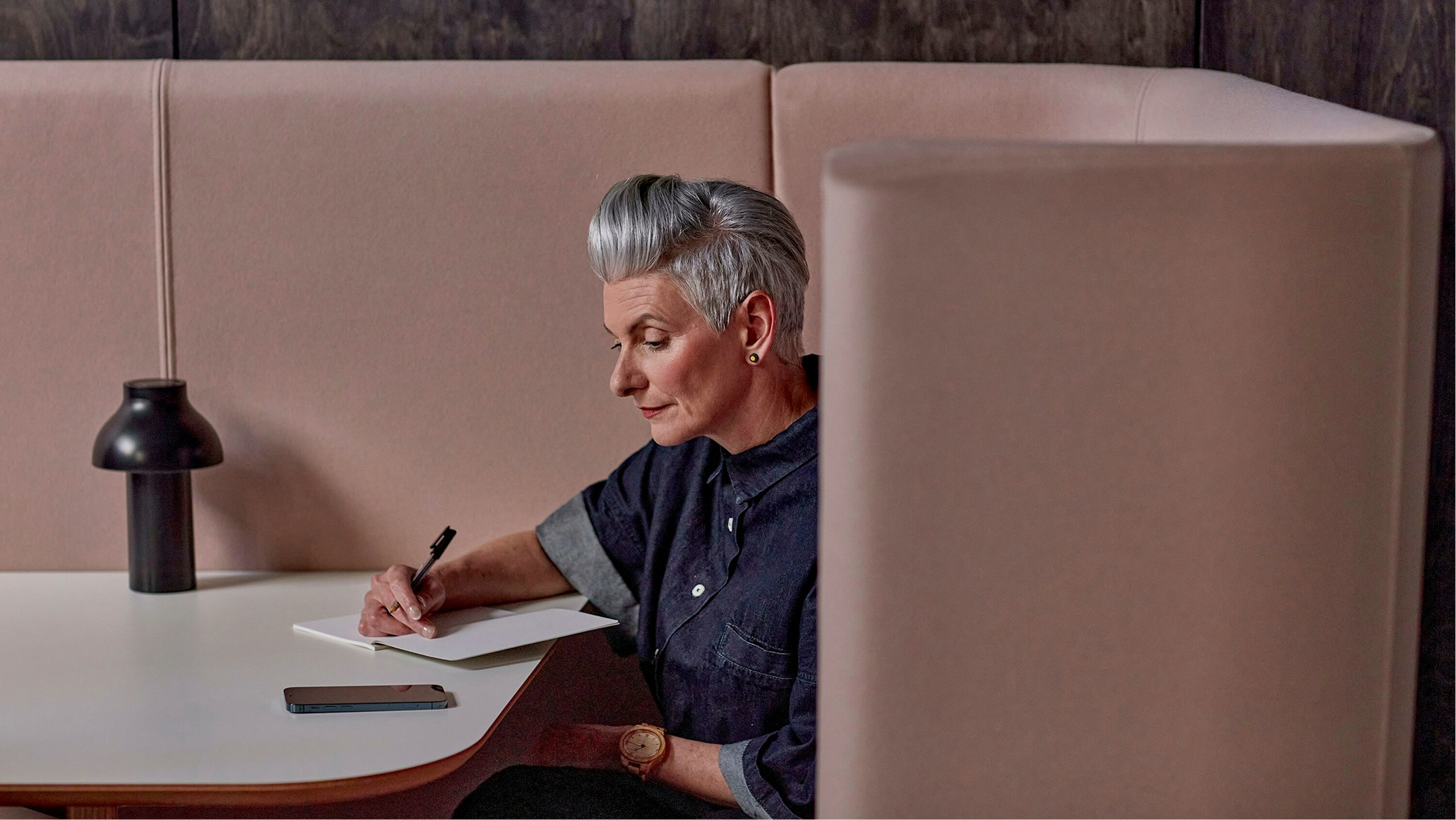 A woman with short grey hair sitting in a light pink booth at a white table while writing in a notebook.