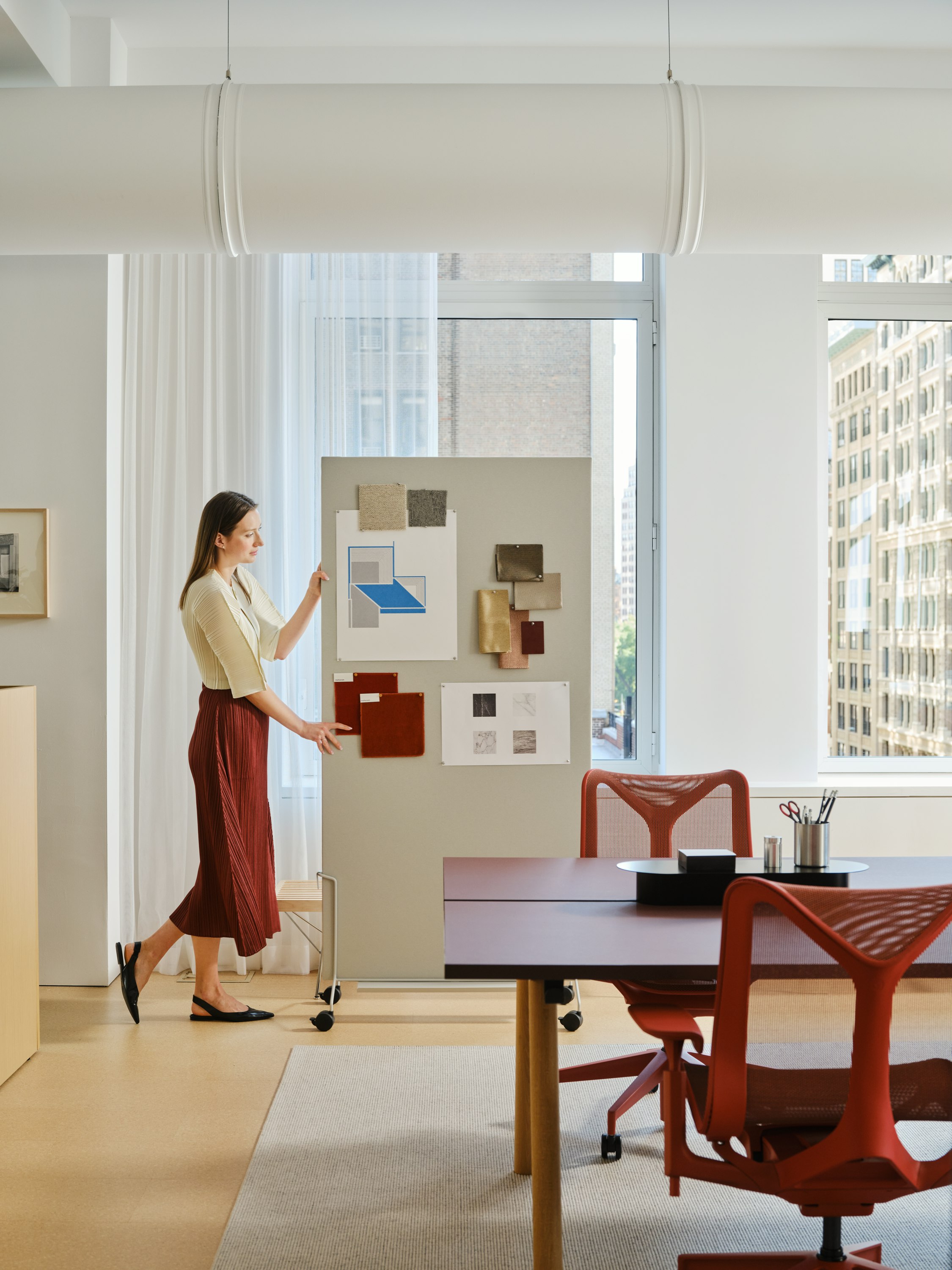  A woman in a red skirt moving an OE1 mobile easel through an office-space furnished with red Herman Miller Cosm Chairs and other furniture from the MillerKnoll collective of brands.