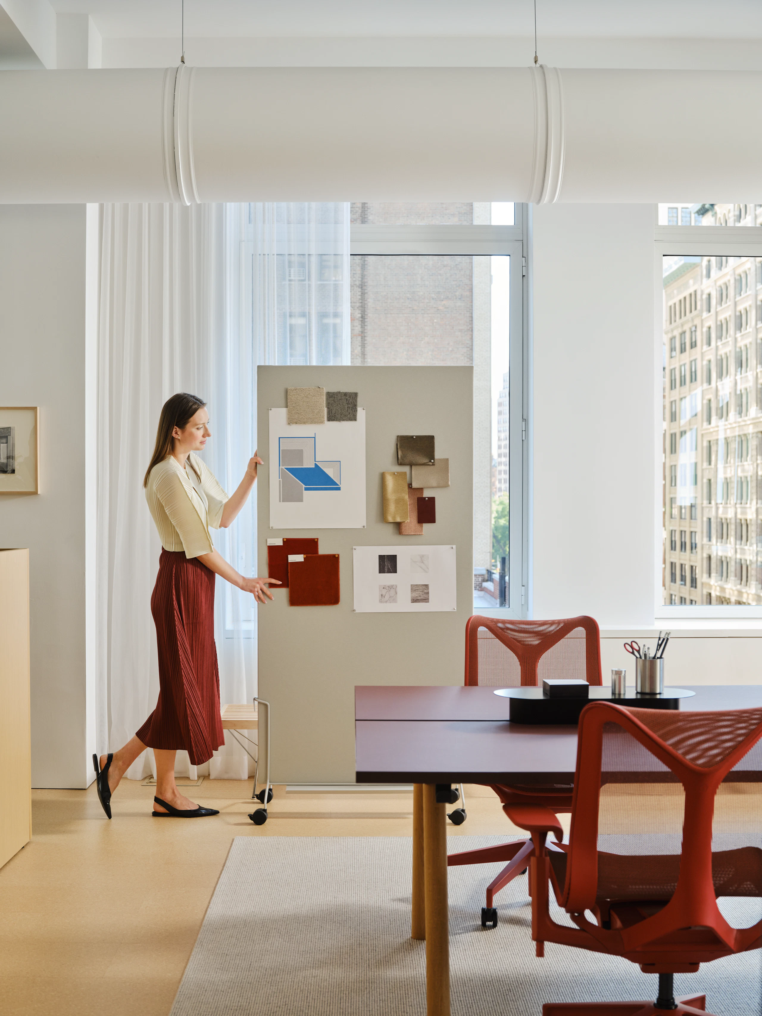 A woman in a red skirt moving an OE1 mobile easel through an office-space furnished with red Herman Miller Cosm Chairs and other furniture from the MillerKnoll collective of brands.