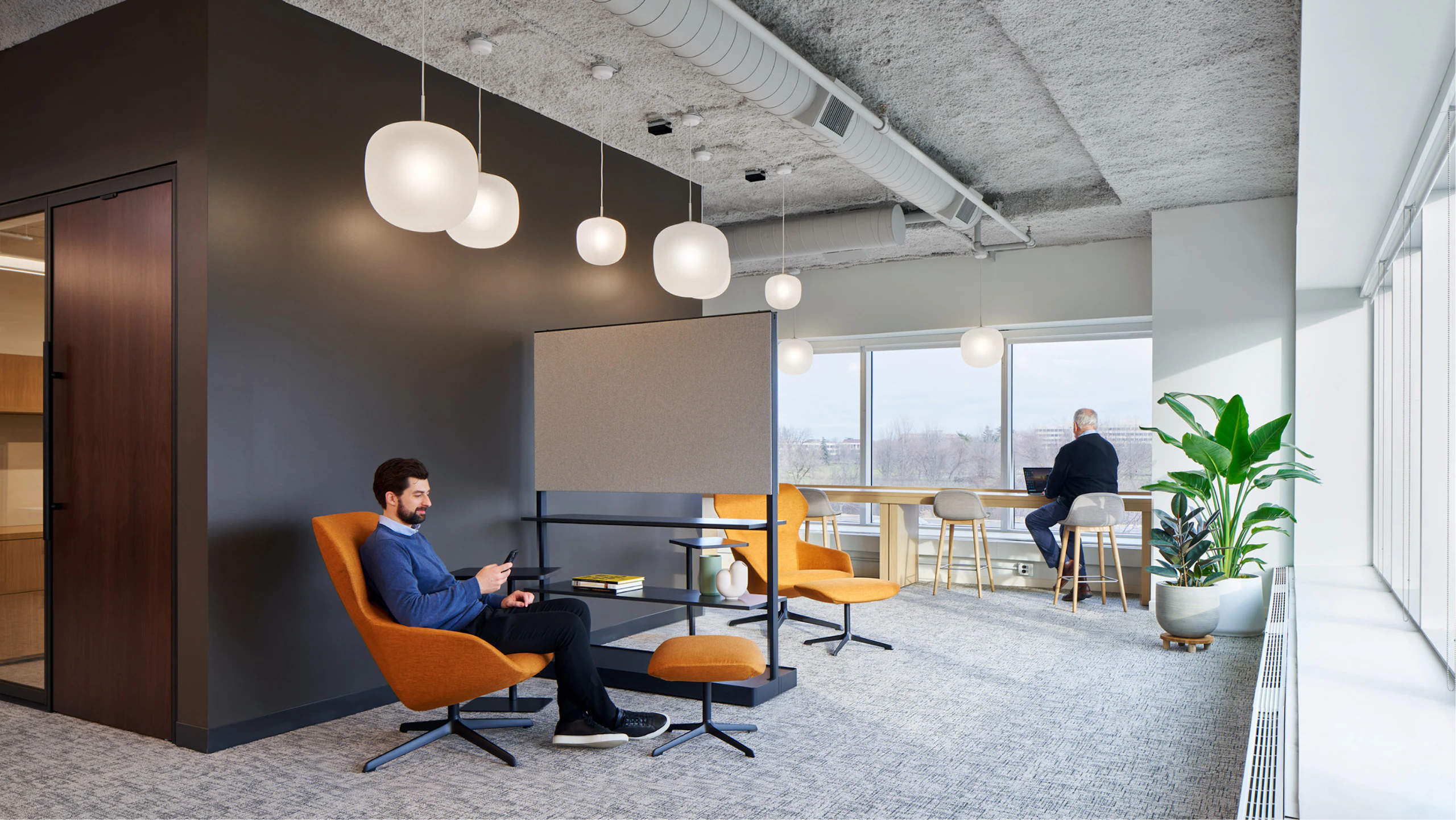 A person in a blue sweater sitting in an orange lounge chair and ottoman while another person works at a bar height counter in the office of an industrial organization