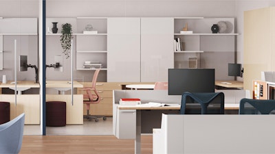 Modern interior with workstations separated by glass panels. Each station features a desk, computer monitor, and chair. Storage units and shelving line the walls, displaying books and decorative items.
