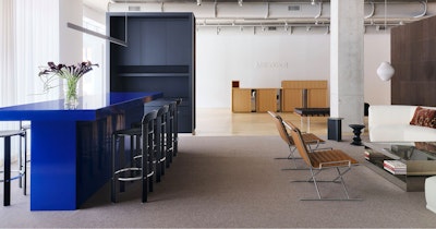 A large, bright blue counter height table with black counter height stools next to a communal seating area with a large white sofa at the MillerKnoll welcome area at Design Days