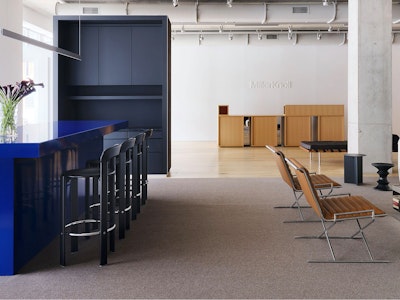 A large, bright blue counter height table with black counter height stools next to a communal seating area with a large white sofa at the MillerKnoll welcome area at Design Days