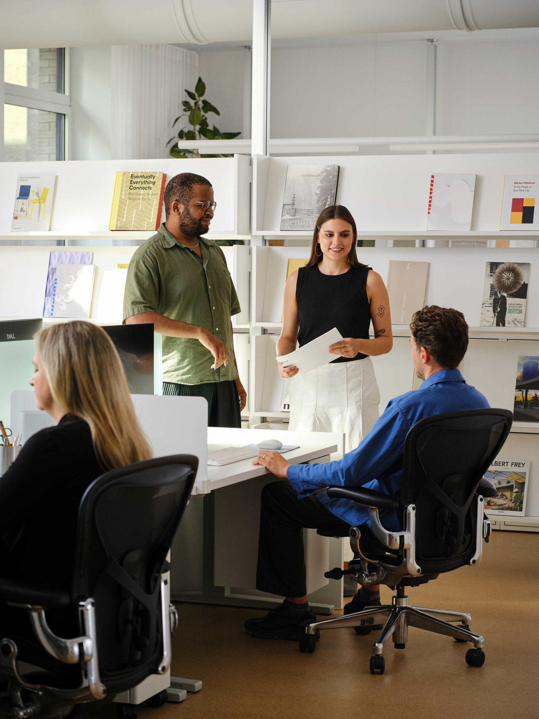 Several people having a conversation around an individual workstation furnished with Herman Miller Aeron Chairs and sit-to-stand desks.