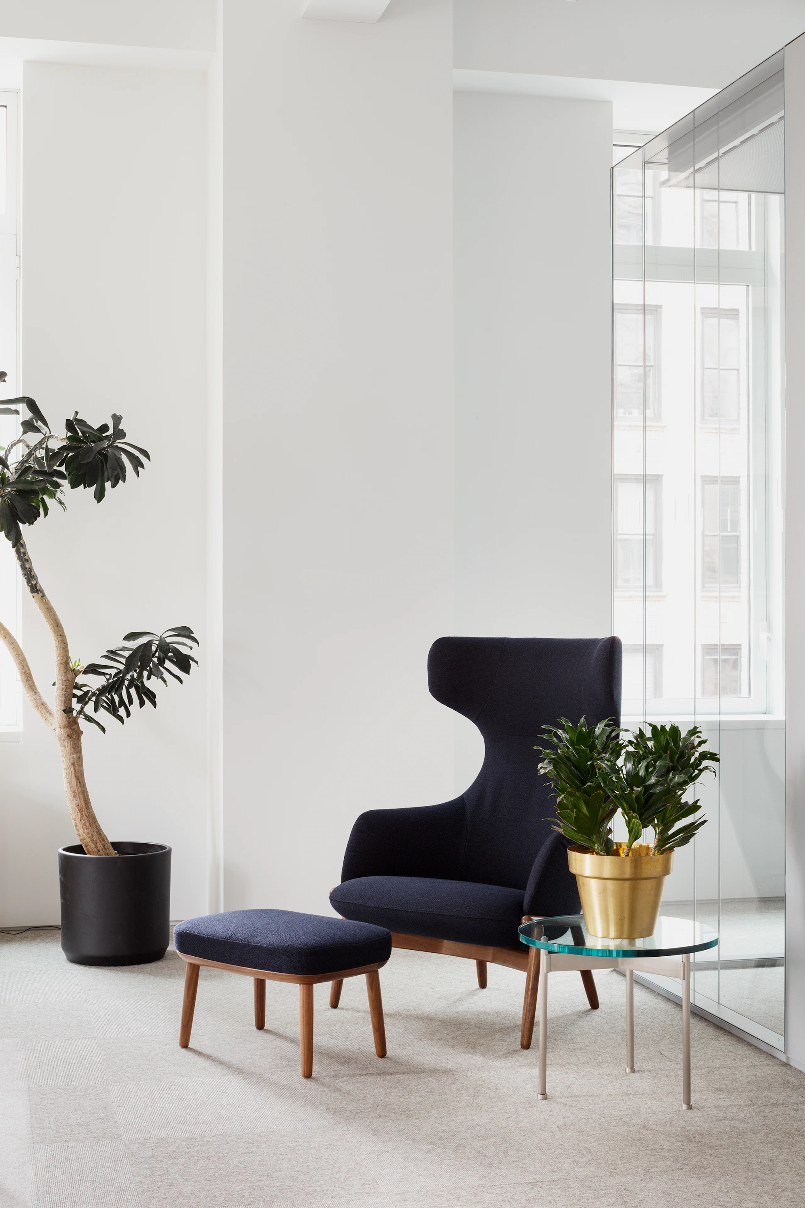 Eye level view of the Reframe Wingback Chair and Crosshatch Ottoman in dark blue with glass round side table with silver metal legs.