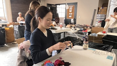 A woman sitting at a table sorting multi-colored pieces of scrap fabric. Others can be seen working next to large bins in the background.