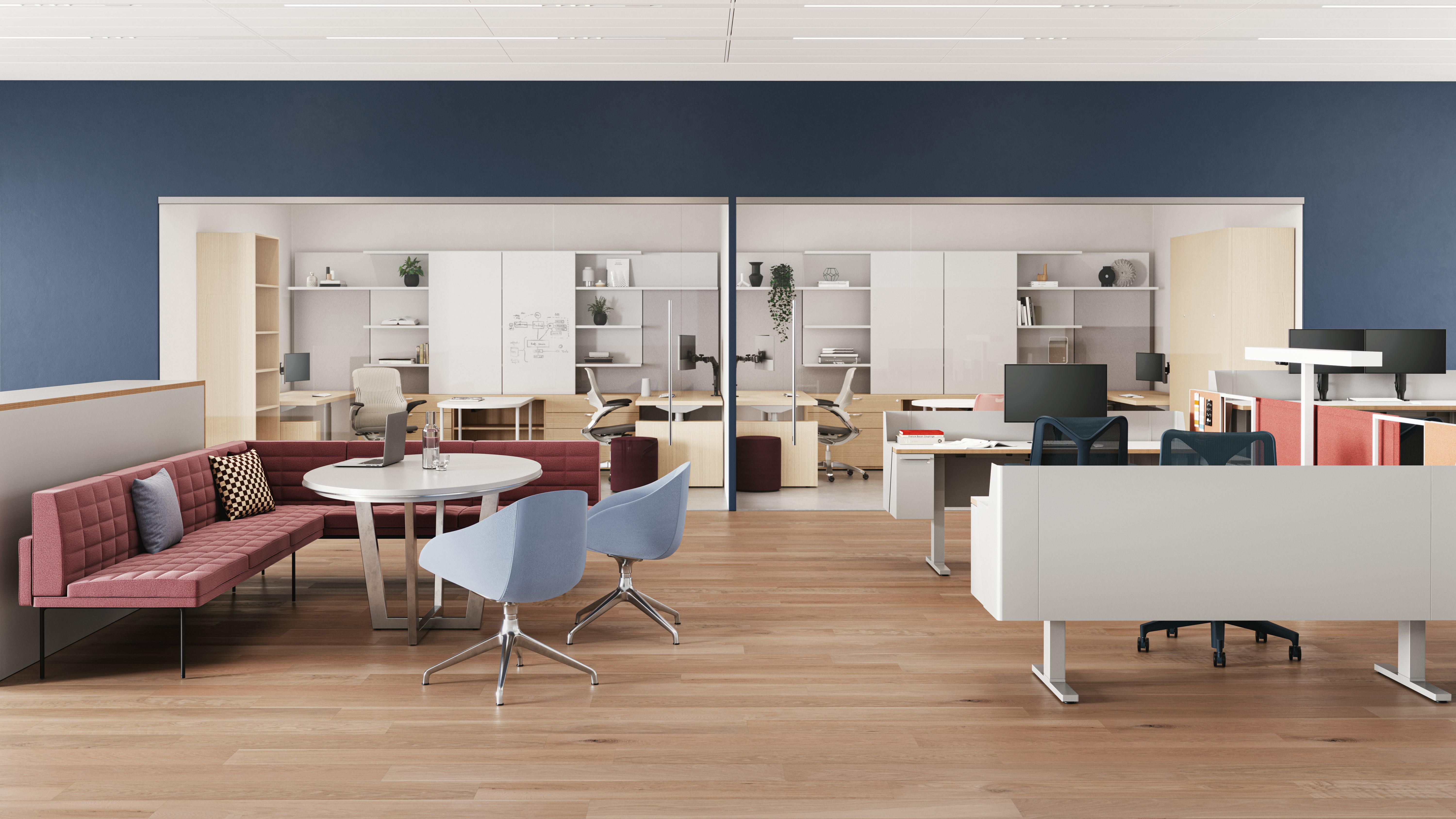 Meeting area with a pink quilted sofa, a round white table, and two light blue chairs. A laptop and water glass are on the table. Workstations visible in the background.