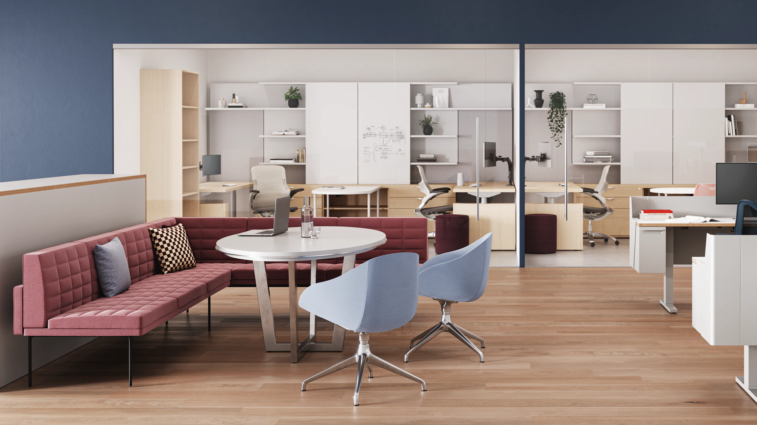 Meeting area with a pink quilted sofa, a round white table, and two light blue chairs. A laptop and water glass are on the table. Workstations visible in the background.