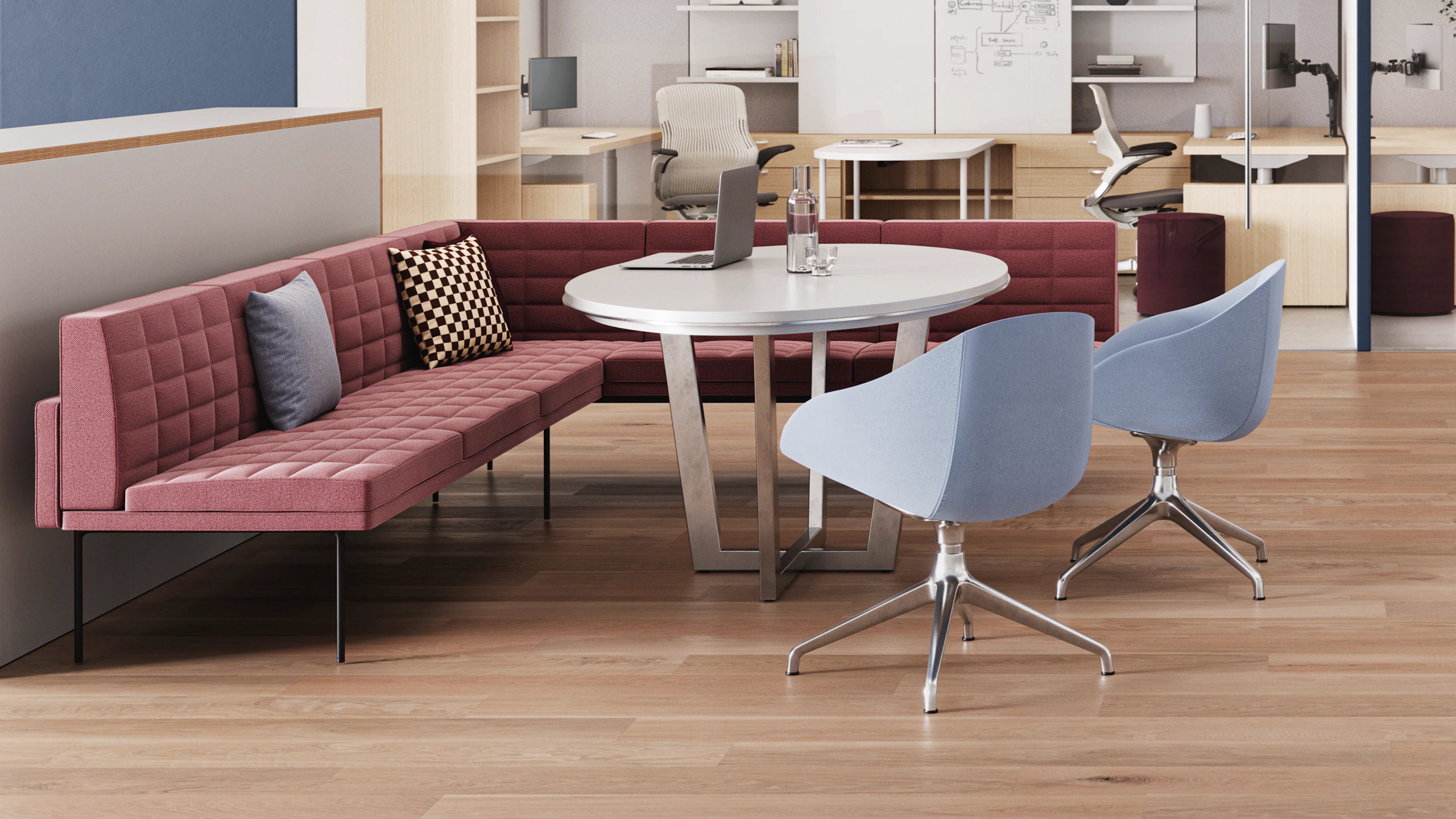 Meeting area with a pink quilted sofa, a round white table, and two light blue chairs. A laptop and water glass are on the table. Workstations visible in the background.