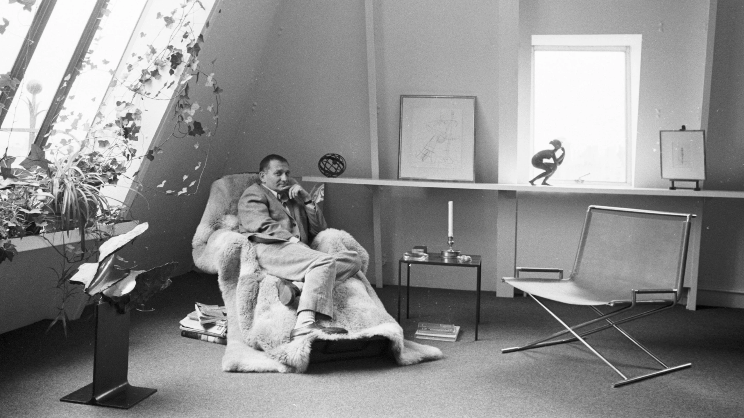 Ward Bennet sitting in a fur covered chair next to large windows, a side table, and several pieces of art.