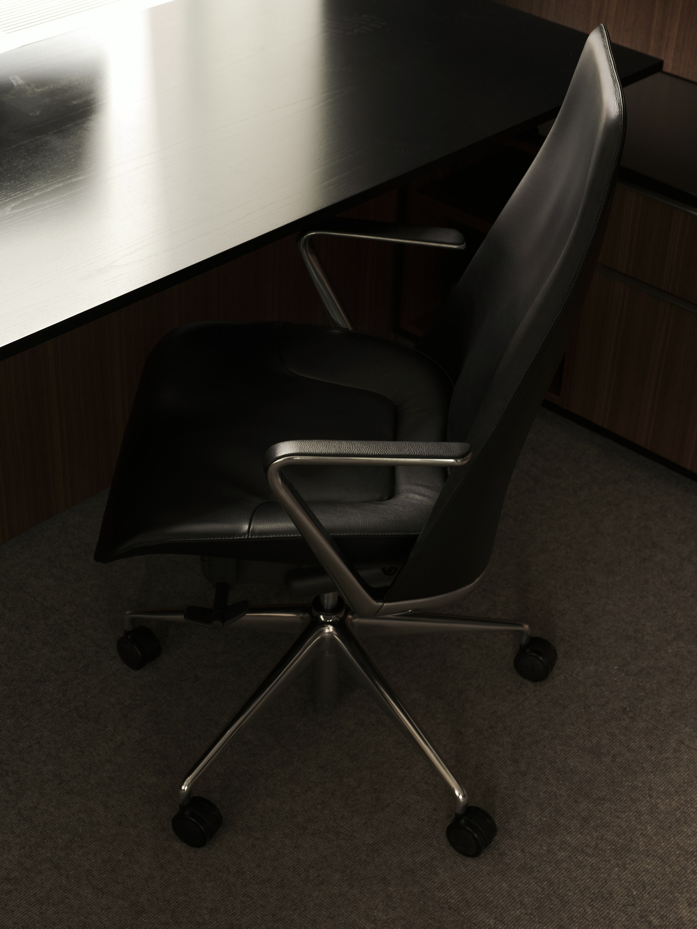 Taper Chair upholstered with black leather with a polished aluminum base next to a work surface in Ebonized Flat Cut Ash, viewed from above at an angle.