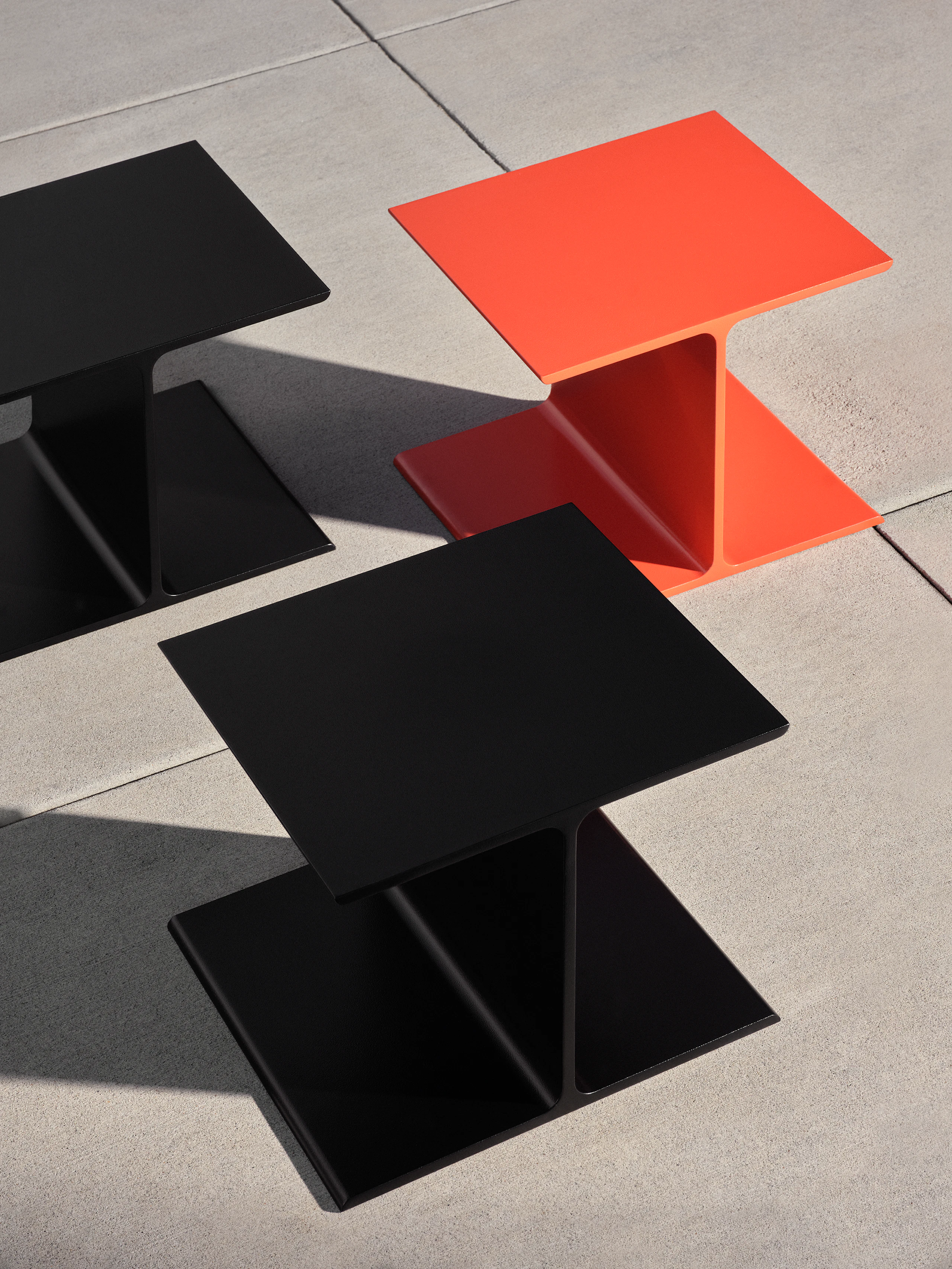 Three I Beam Side Tables designed by Ward Bennett, one in International Orange and two in black, viewed from above at an angle on a concrete floor.