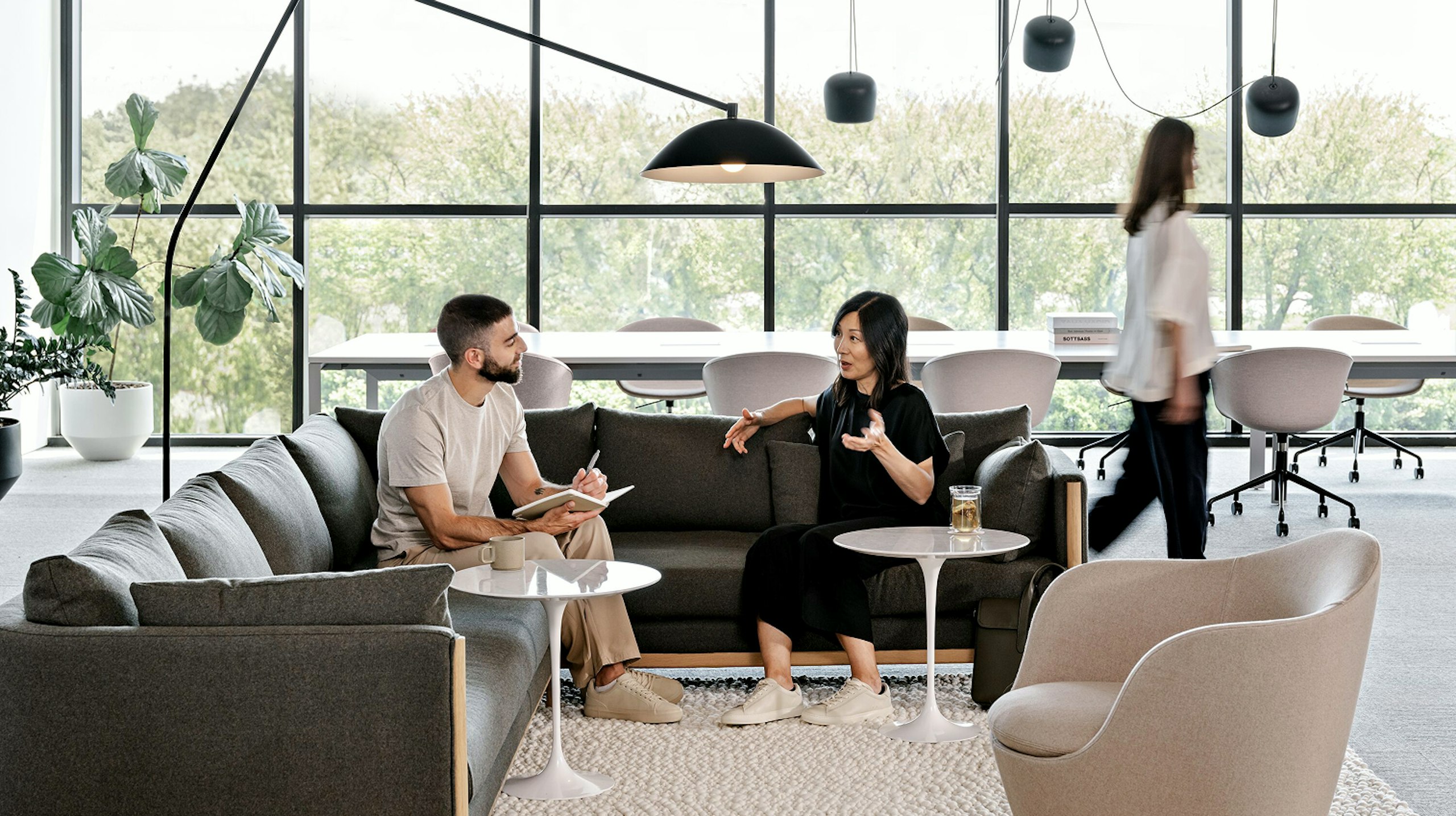 Two people having a conversation on a dark grey L-shaped couch next to white Saarinen side tables. A person in the background walks past windows and a large conference table and task chairs.