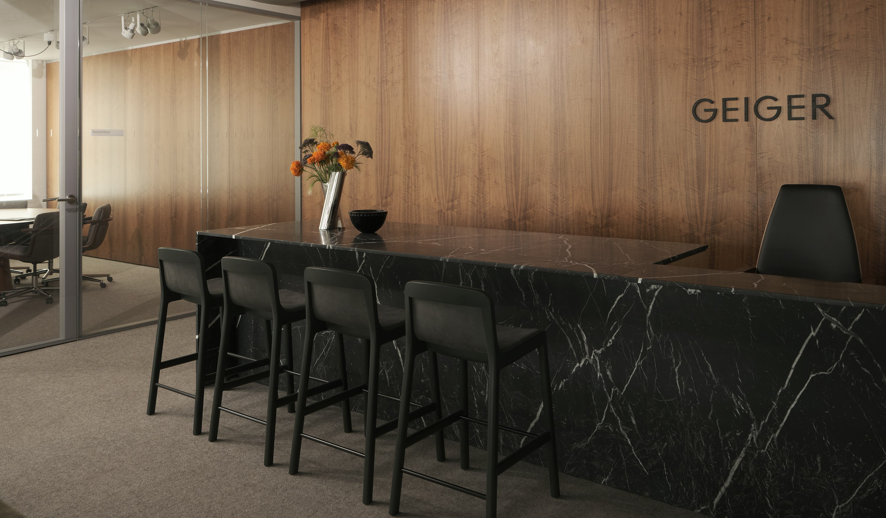 A custom Geiger reception desk in Nero Marquina Marble with four counter height Lijn Stools in Ivy and a Clamshell Chair against a veneer wall in the Geiger Chicago showroom, viewed at an angle.