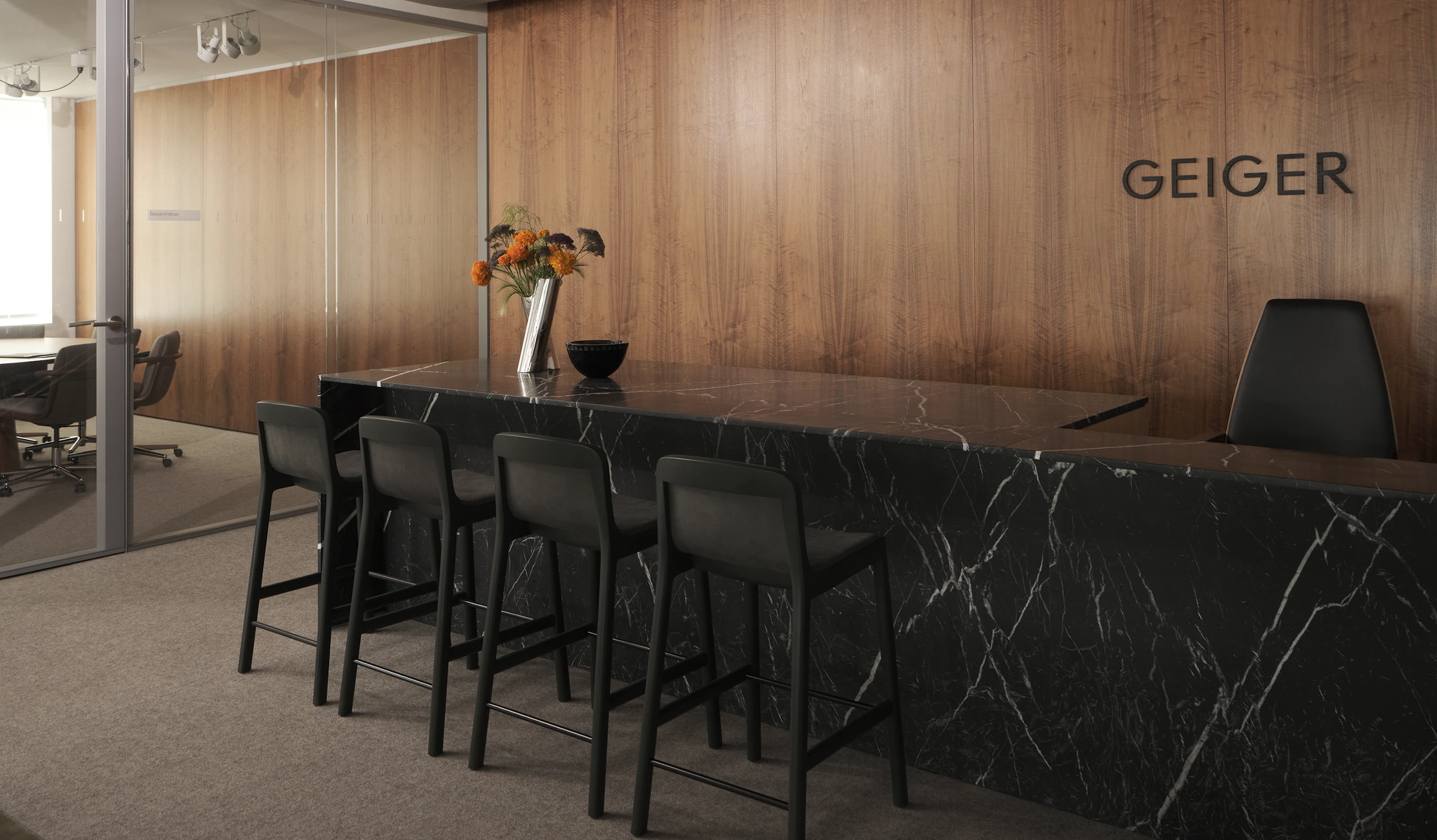 A custom Geiger reception desk in Nero Marquina Marble with four counter height Lijn Stools in Ivy and a Clamshell Chair against a veneer wall in the Geiger Chicago showroom, viewed at an angle.