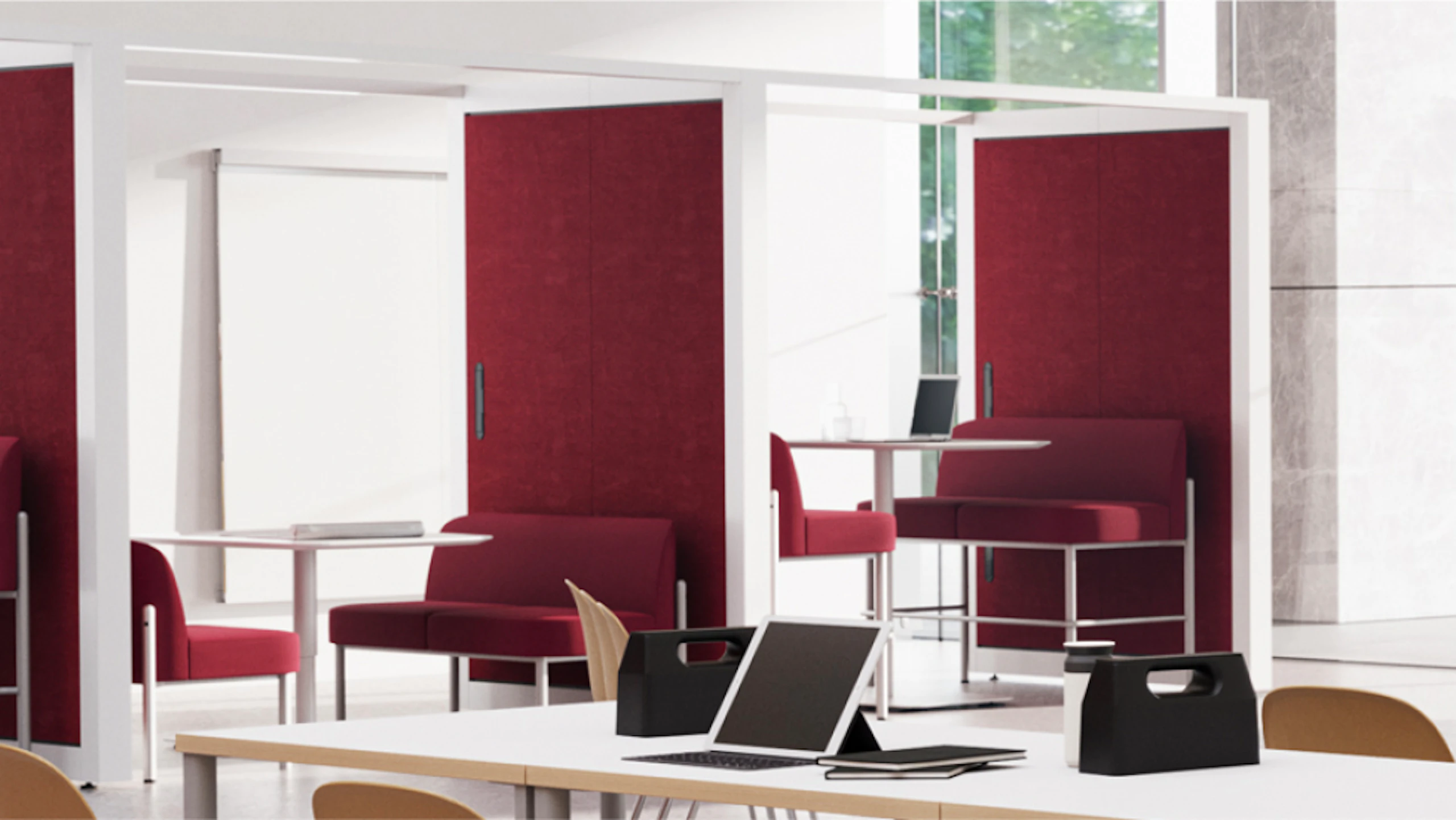 Modern booths with red fabric panels and white frames. Some booths have small tables and seating inside. A long white table in the foreground has laptops, bags, and notebooks on it.