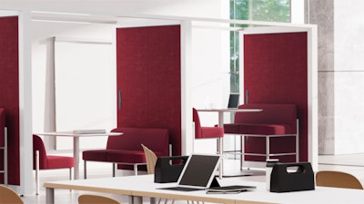 Stands modernos con paneles de tela roja y marcos blancos. Algunos stands cuentan con pequeñas mesas y asientos en su interior. En primer plano hay una mesa blanca larga con ordenadores portátiles, bolsos y cuadernos.