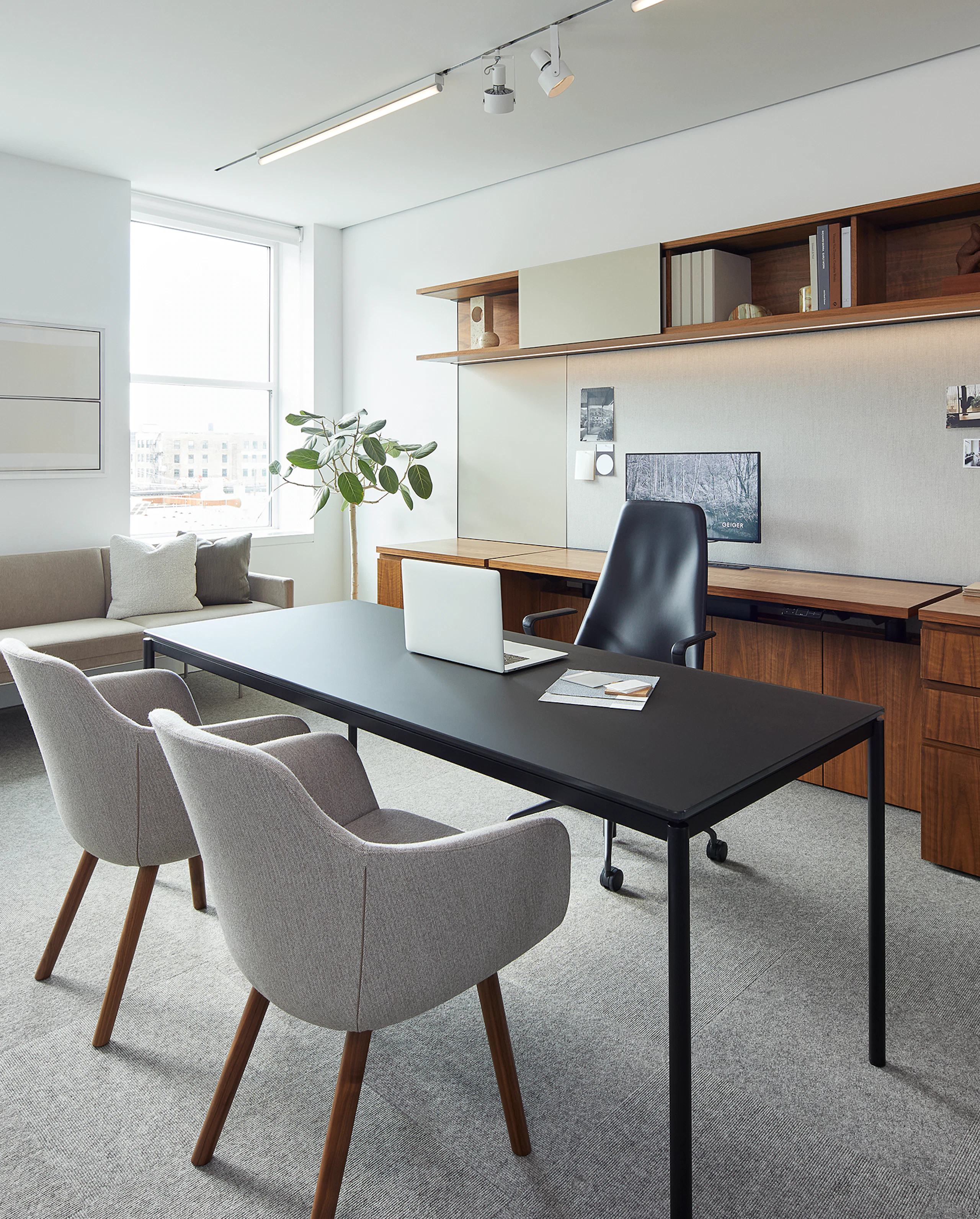 A black desk with two grey guest chairs in front of it sit in a spacious private office with dark wood shelves and storage in the background.