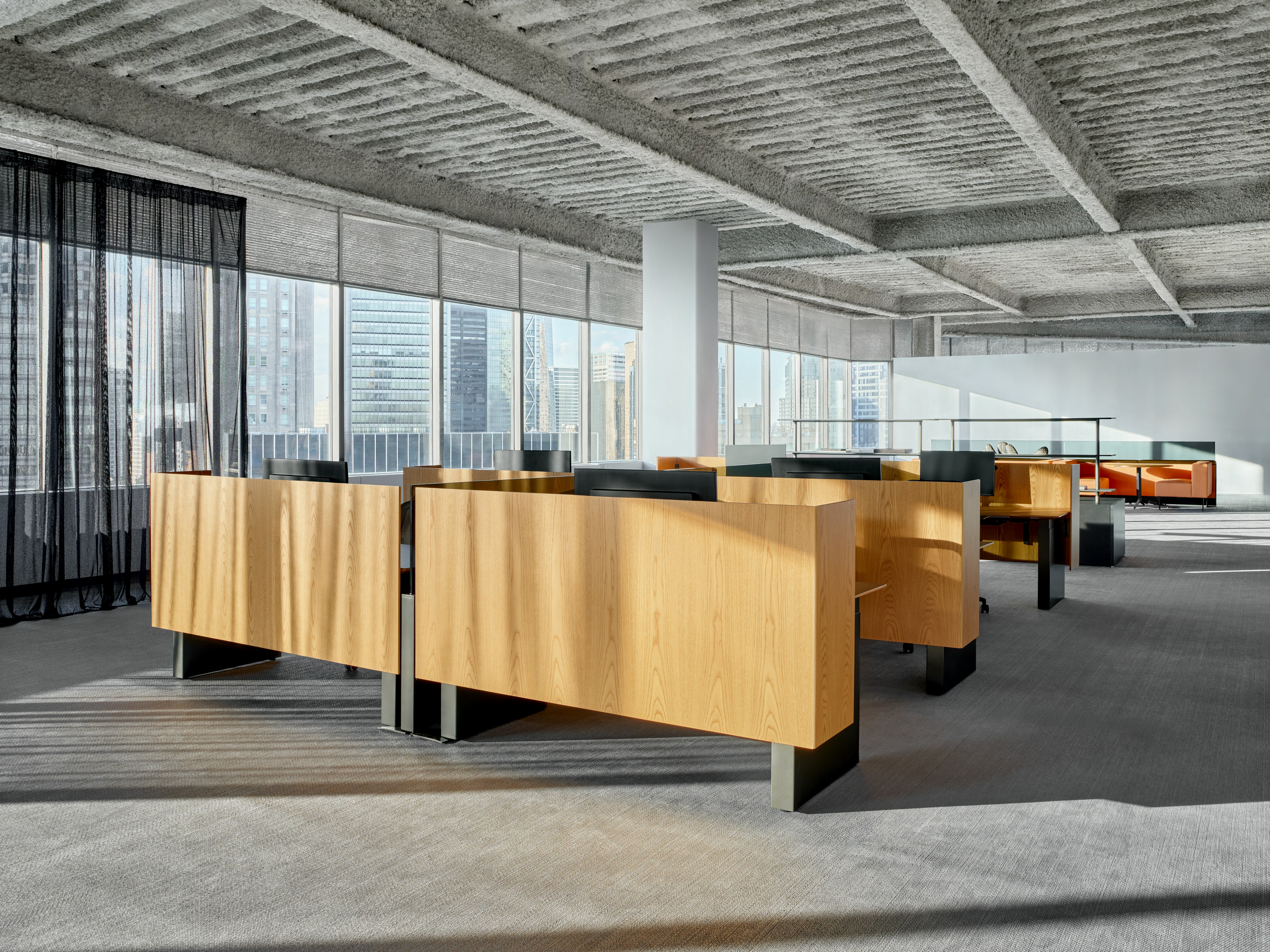 A group of workstations are in an open office space in front of a large wall of windows. Through the windows the New York City Skyline is visible. Behind the workstations is a shared storage space as well as some casual meeting spaces.