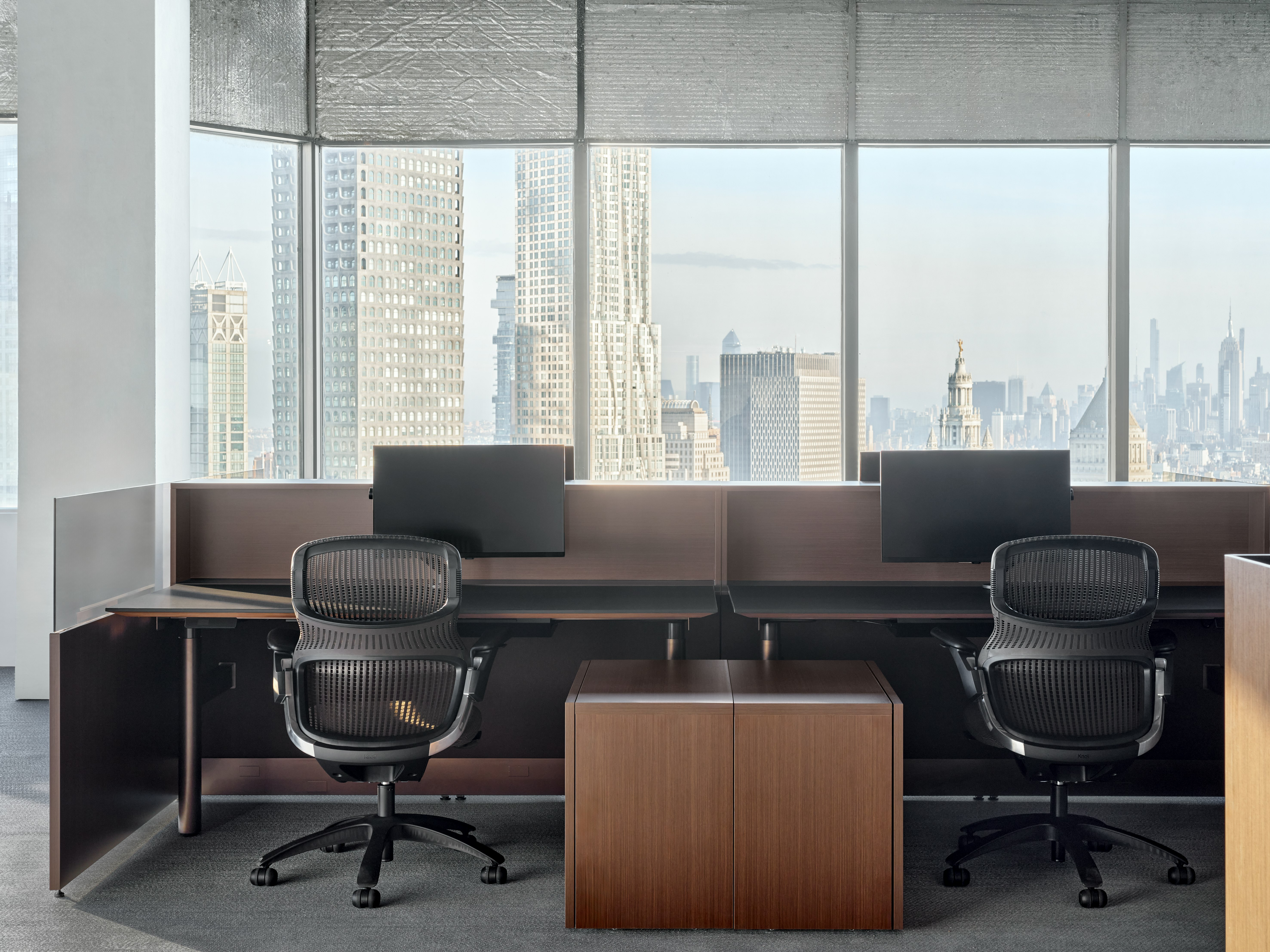 A group of panel based workstations are in front of a large group of windows that show the New York City Skyline. At each workstation is a height adjustable table, a work chair and a personal storage pedestal.
