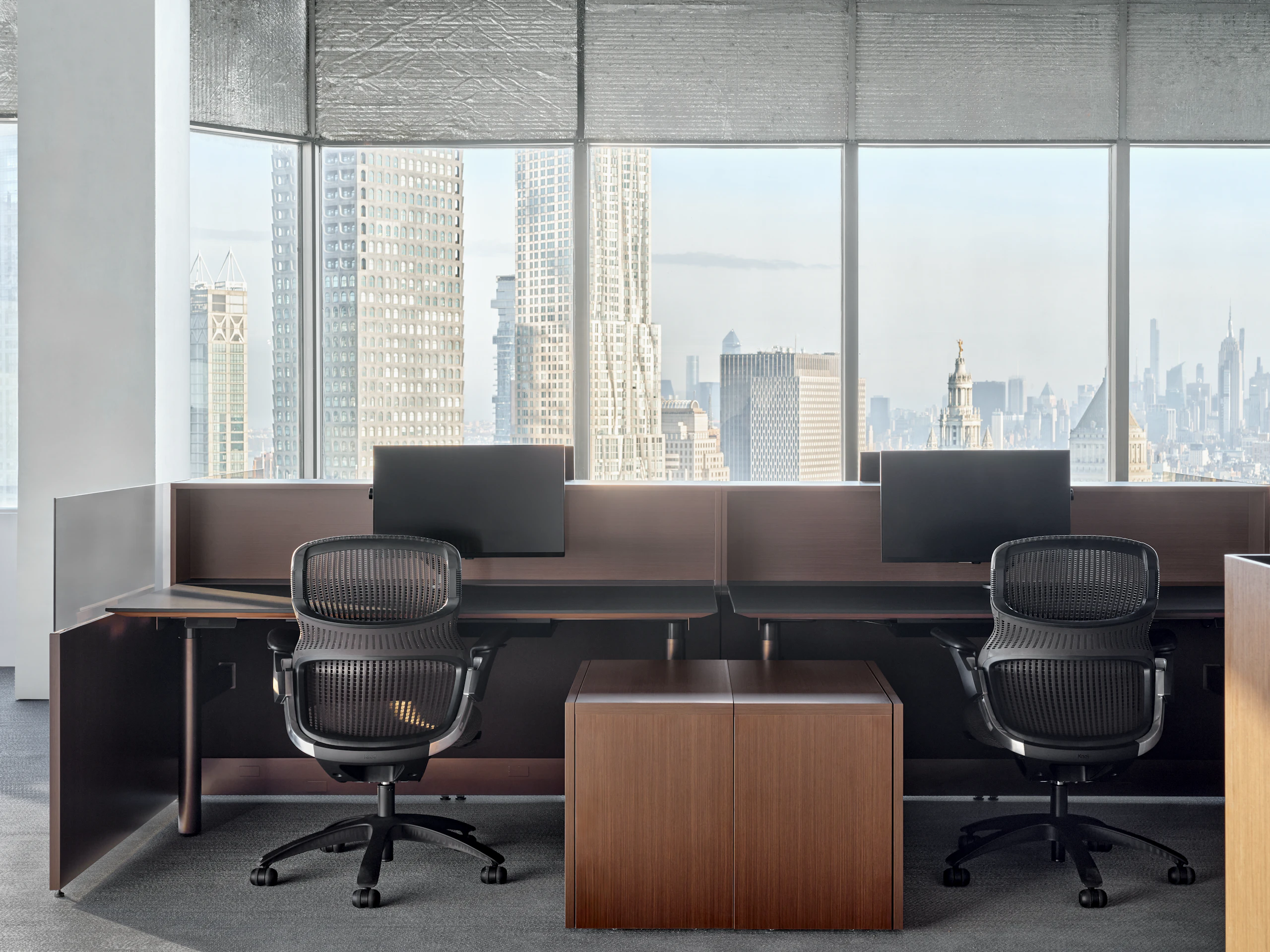 A group of panel based workstations are in front of a large group of windows that show the New York City Skyline. At each workstation is a height adjustable table, a work chair and a personal storage pedestal.