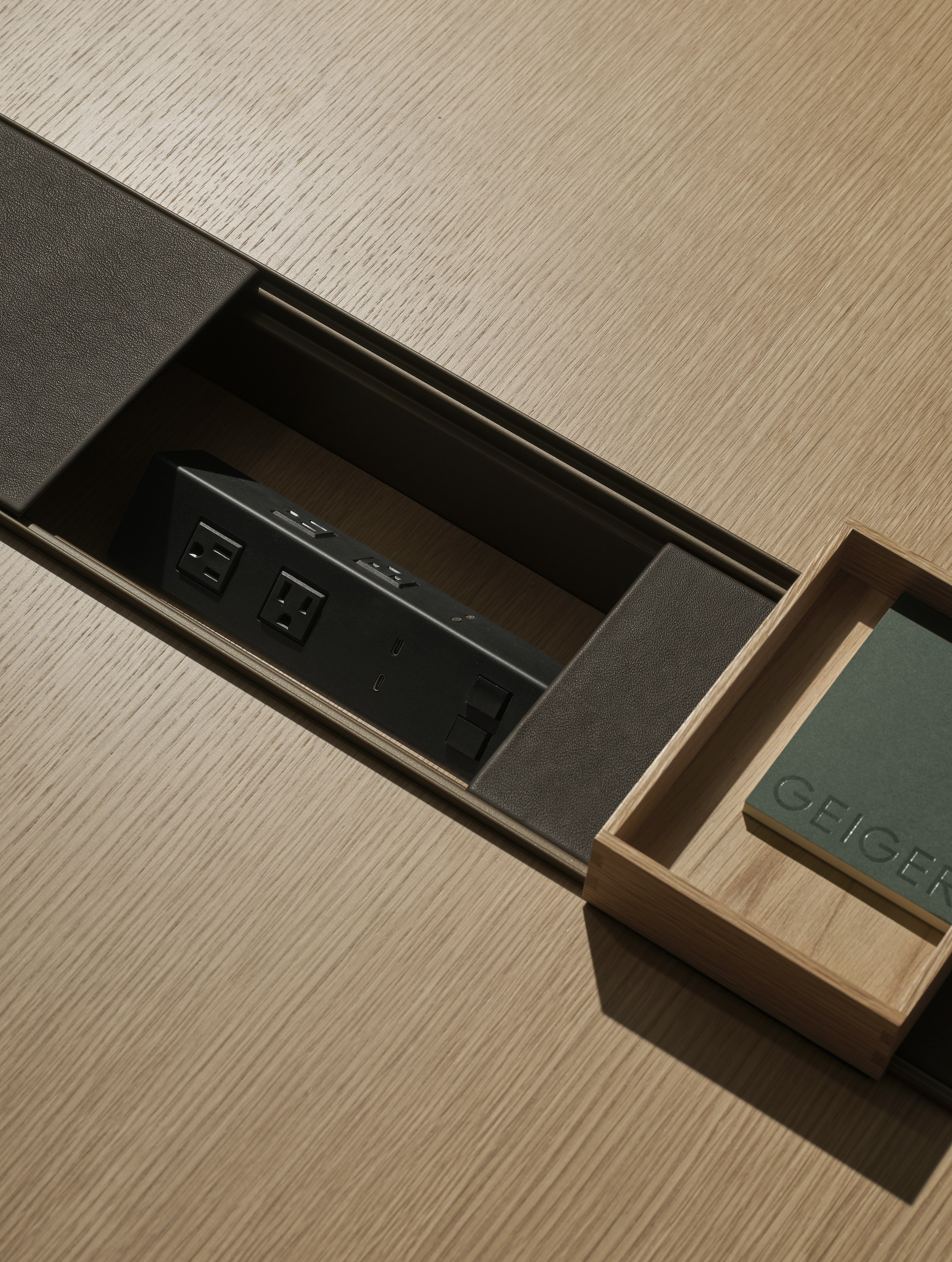 Detail view of an open Ascent Conference Table power trough with leather covers and an accessory tray containing a Geiger notebook, with a Natural Rift Cut Oak tabletop, viewed from above at an angle.