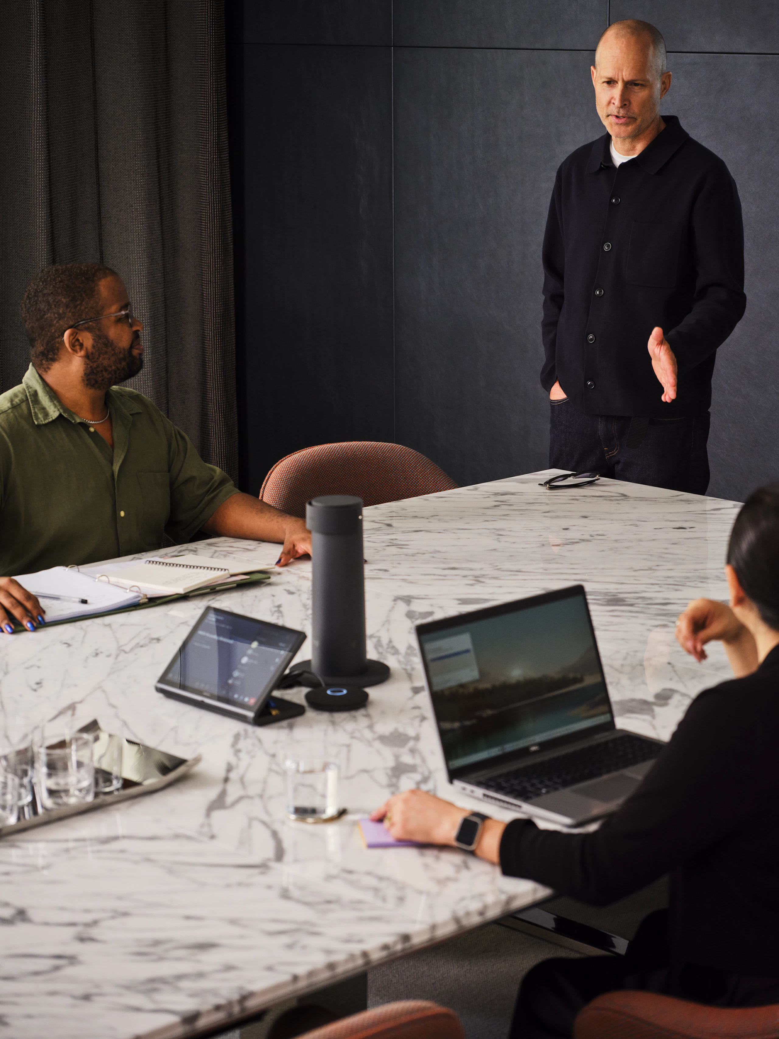 Three people, two seated with laptops and one standing, having a meeting at a large conference table.