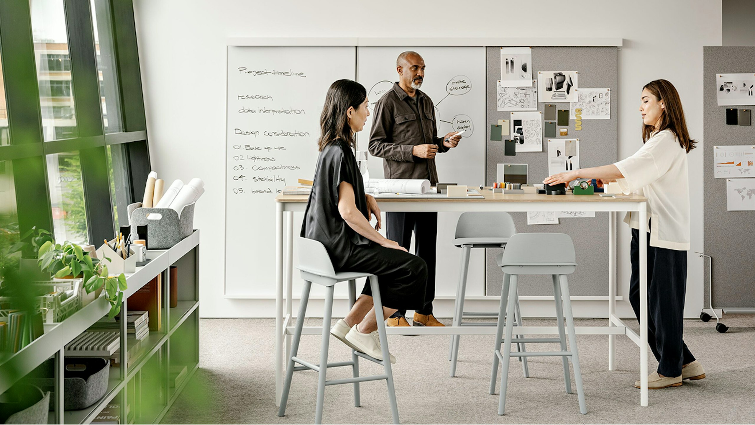 Two women and one man stand around a table while working in front of a whiteboard. Various objects and pieces of paper can be seen on the table and pinned to the board.