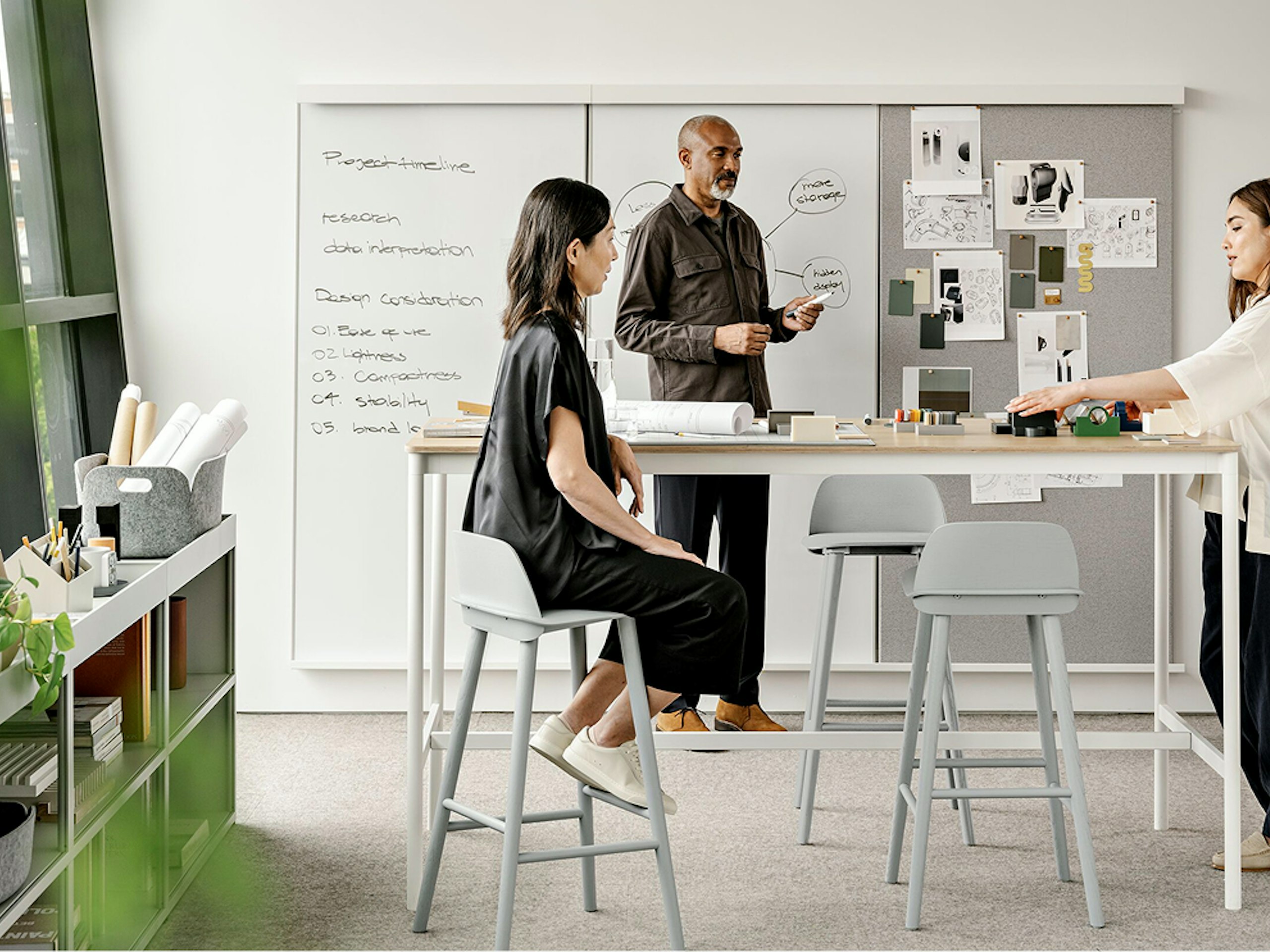 Two women and one man stand around a table while working in front of a whiteboard. Various objects and pieces of paper can be seen on the table and pinned to the board.