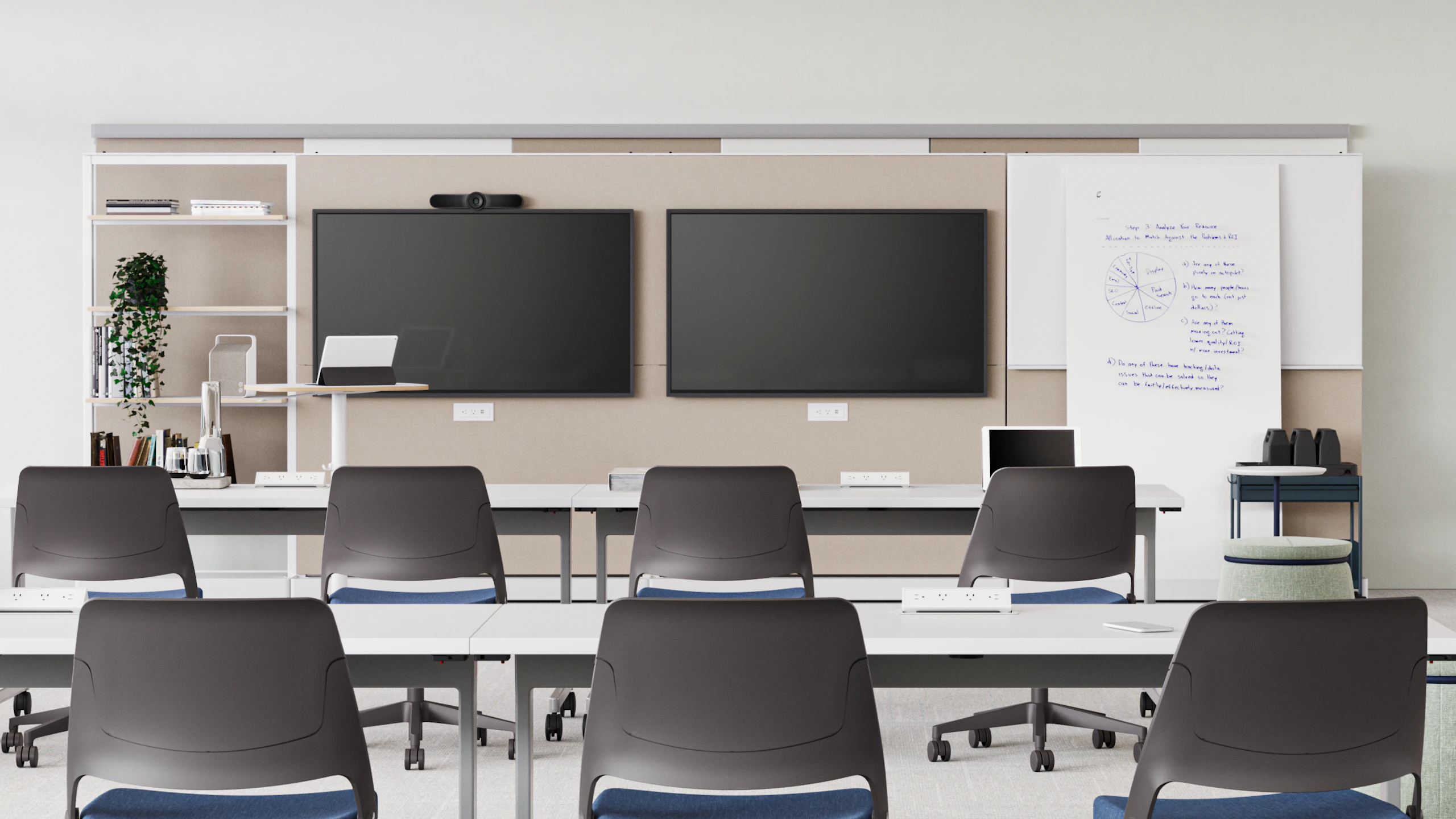 Modern classroom room with two large screens, a whiteboard with notes, and a long white table surrounded by teal chairs. Shelving unit with books and plants in the background.