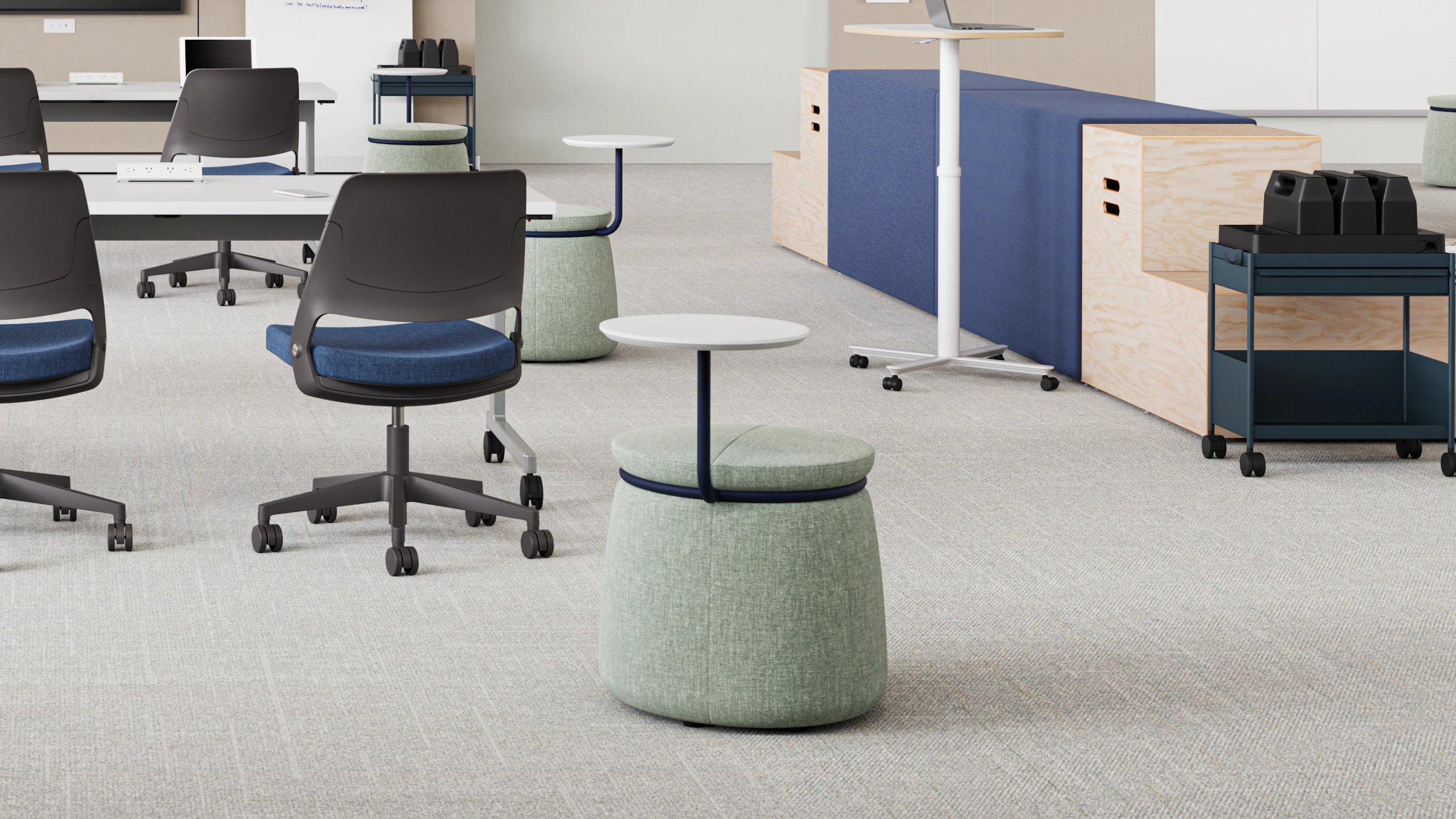 Modern classroom featuring a long white table with teal chairs, and unique mobile stools with small, attached desks. Modular storage and a standing desk are in the background.