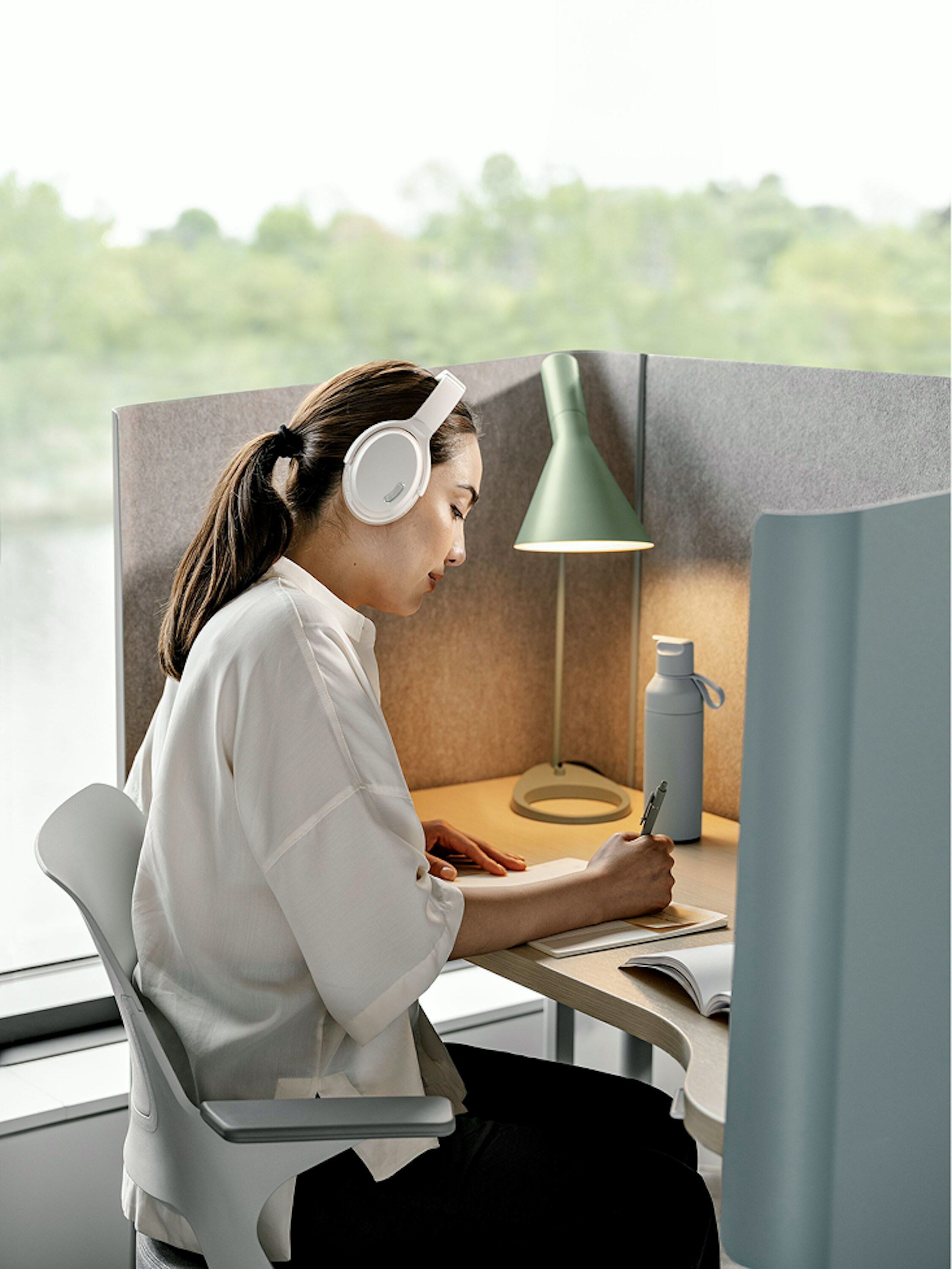 A person with a long brown ponytail, wearing a white shirt and white headphones, sitting in a white and grey chair while writing on a notepad at a private workstation.