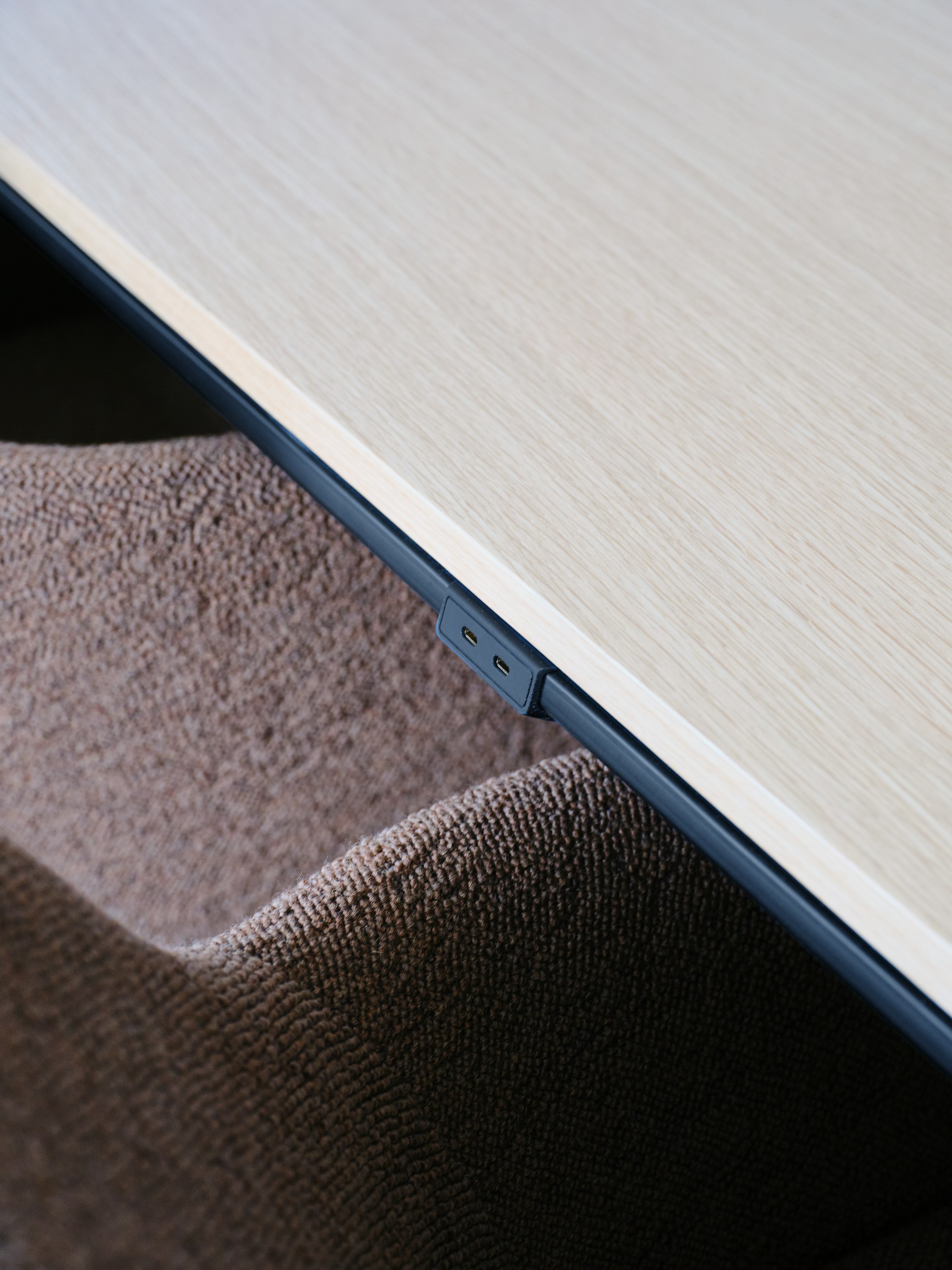 Detail view of the Ascent Conference Table edge power in black with a Natural Rift Cut Oak tabletop and a Saiba chair upholstered in a brown textile, viewed at an angle.