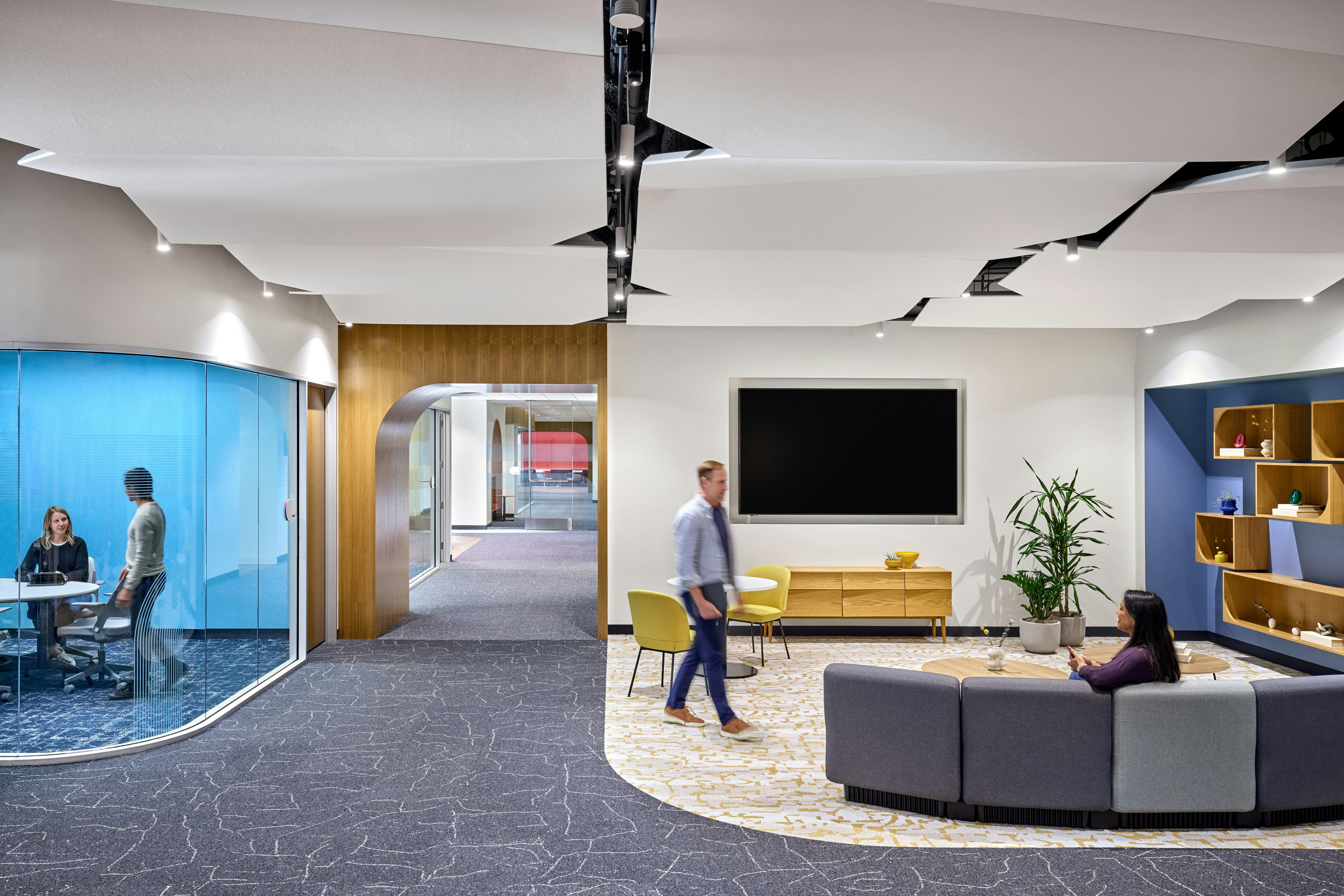 Contemporary office setting with modular lounge seating, coffee and cafe tables, wall-mounted shelving, and an adjacent meeting room with curved glass enclosure.