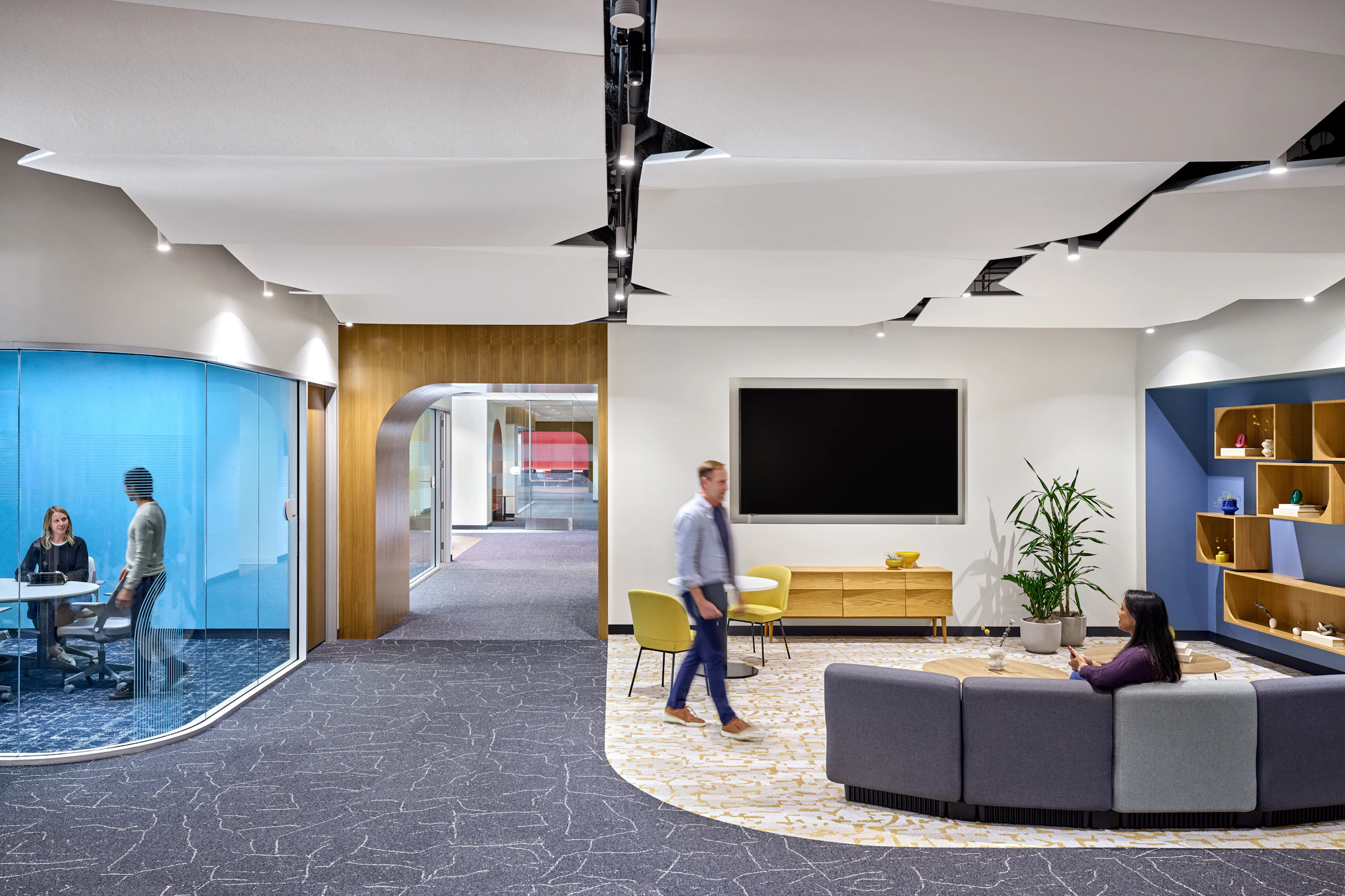 Contemporary office setting with modular lounge seating, coffee and cafe tables, wall-mounted shelving, and an adjacent meeting room with curved glass enclosure.