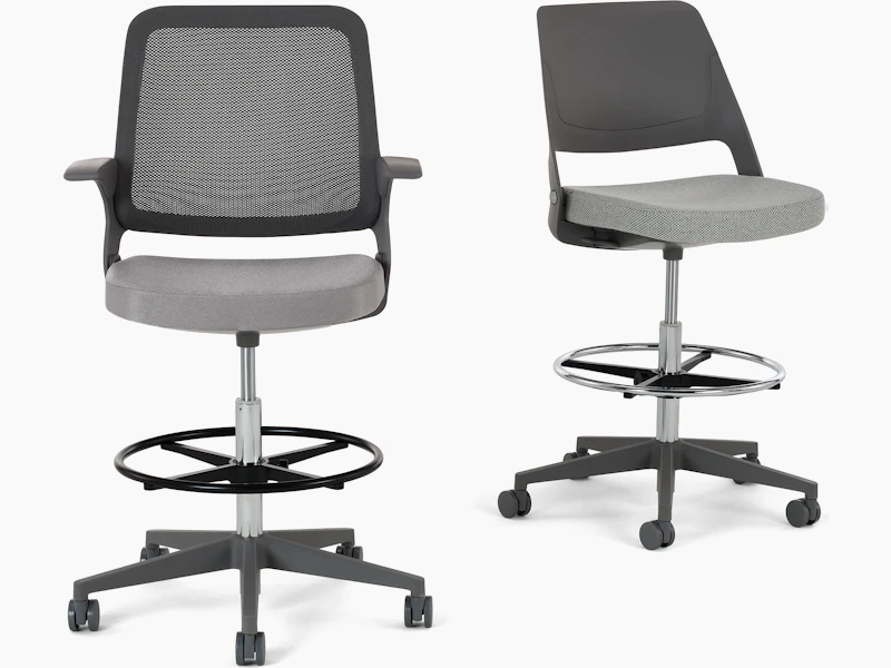 Ollo High Task Chair Stool with a Gunmetal color knit back, medium grey frame and base, fixed arms, a 5-star plastic base, and hard casters, with a silver cylinder and a silver foot ring. The cushioned seat is upholstered in Reflect textile in the color 4 Steel (grey). Next to an Ollo High Task Chair Stool with a medium grey shell back and base, no arms, a 5-star plastic base, and hard casters, with a silver cylinder and a silver foot ring. The cushioned seat is upholstered in Facet textile in the color 2 Mica (light grey). White background. White baground